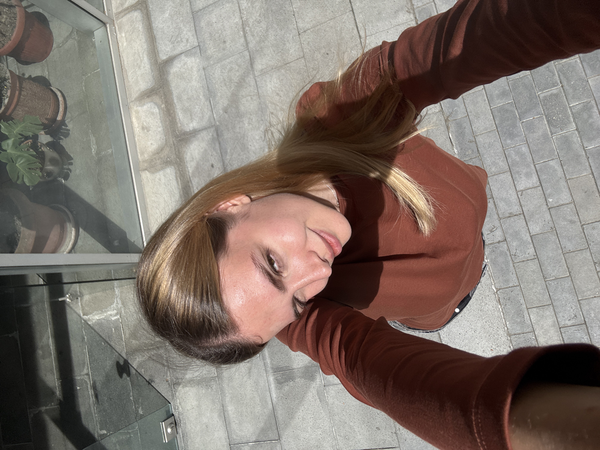A woman takes a selfie outdoors in Tbilisi, Georgia, with gray stone pavers underfoot. She wears a brown long-sleeved top. To her left are potted plants and a glass partition.
