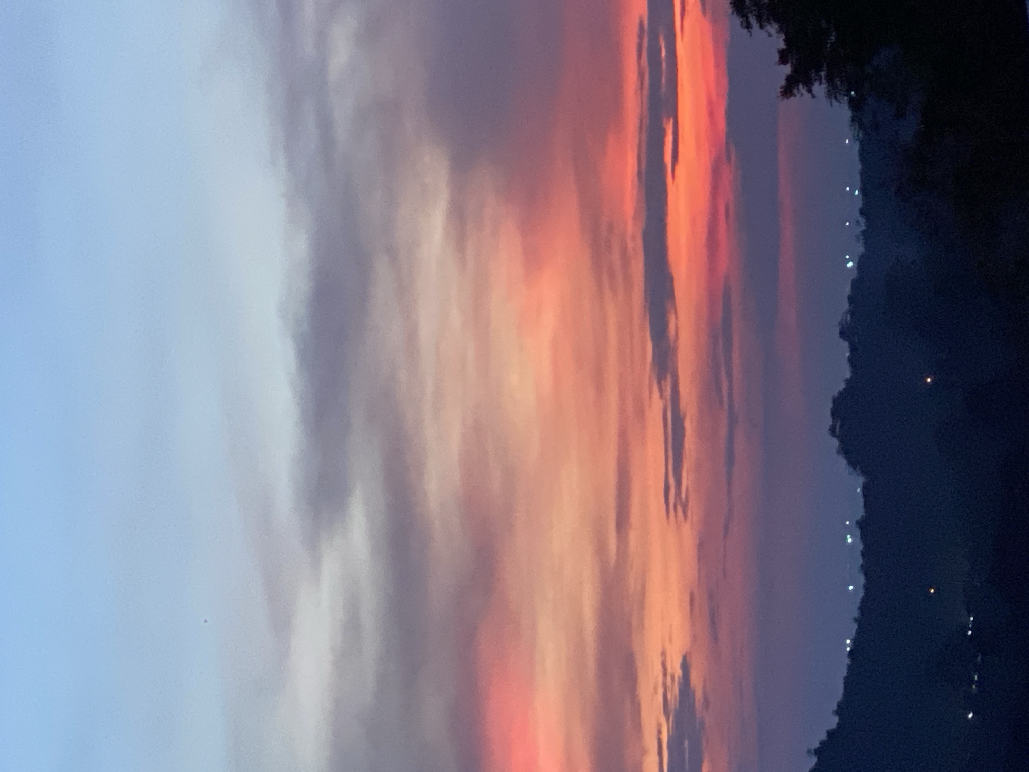 A vertical, full shot depicts a colorful sunset over Baan Don Sai, Thailand. The sky transitions from light blue on the left to orange and red hues on the right, with cloud formations stretching across the frame. The right side of the image shows a dark, silhouetted landmass dotted with faint lights, possibly indicating settlements or streetlights in Baan Don Sai. The foreground details of the city are not discernible due to the darkness. The photograph captures a tranquil evening scene.