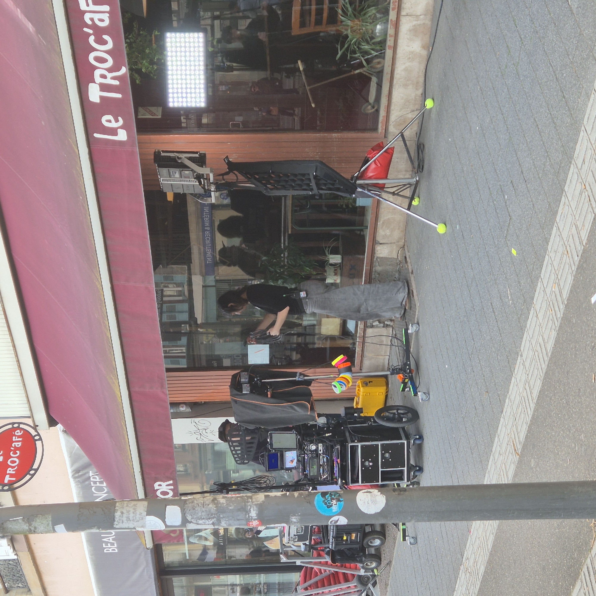 This image depicts an active film or video production setup on a grey paver sidewalk in front of "Le Troc'afé" in Strasbourg, France. The cafe is identifiable by its prominent red awning with "Le TROC'afé" in white lettering and an oval red sign above it. An adjacent storefront to the left displays "BEAU CONCEPT" on a grey awning.
In the mid-ground, a person with dark hair, wearing a black t-shirt and grey wide-leg trousers, stands with her back mostly to the viewer. She is engaged with a wheeled, multi-monitor equipment cart, holding cables, indicating a technician role. To her right, a large rectangular LED light panel on a tripod stand casts bright white light. Another black grid-like panel on a separate tripod, stabilized by a red sandbag and utilizing yellow tennis balls as foot covers, stands nearby. A yellow equipment case and a stand with multiple colored tape rolls are also present on the sidewalk. To the left of a grey utility pole, additional wheeled production carts laden with cases and red sandbags are visible. Black cables are arrayed across the paved surface. Storefront windows reflect interior elements, reversed signage, and indistinct figures.