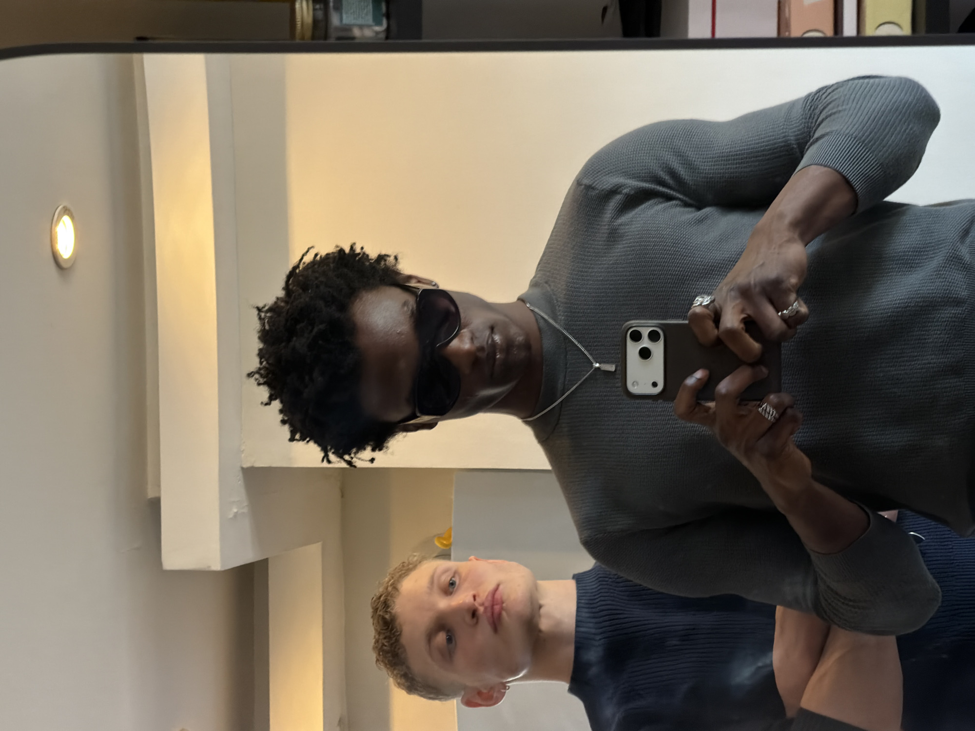 A vertically oriented image shows a reflection in a mirror. A dark-skinned man in sunglasses is holding a phone and taking a photo of himself and another man in the mirror. The first man is wearing a gray textured shirt and a silver chain necklace. He has rings on his fingers. The second man has short blond curly hair and is wearing a dark blue shirt. The background is a white wall with molding and a built-in light. There is a shelf above with various items on it. This image has no indication of being in the city of London.