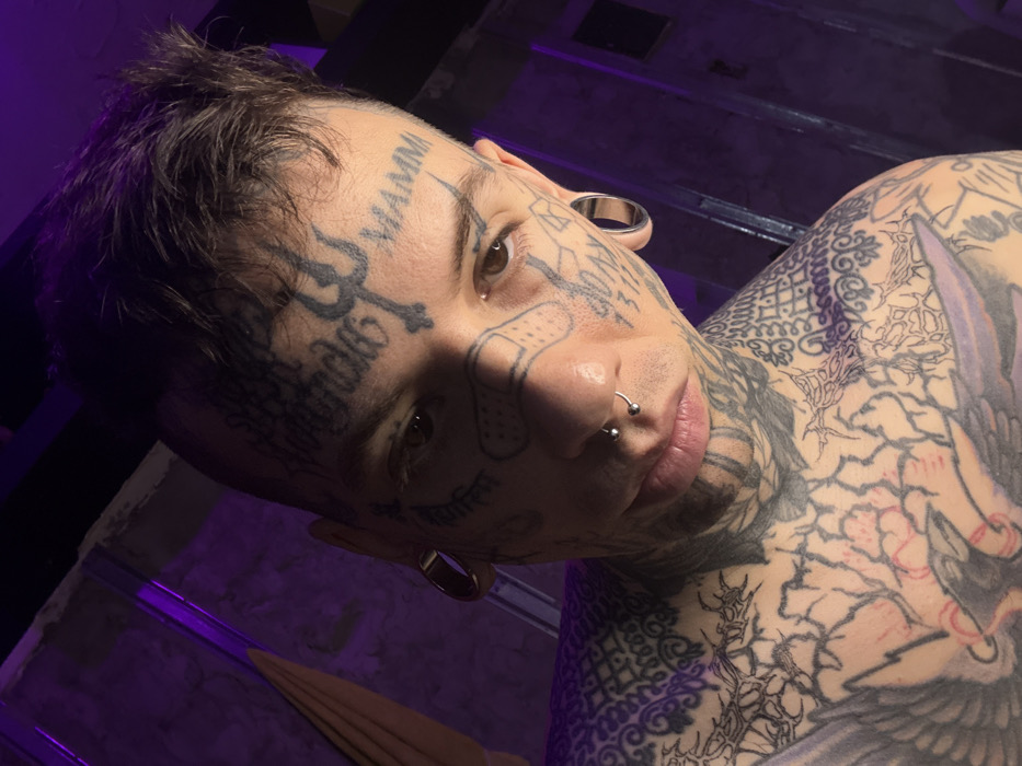 A close-up, slightly high-angle portrait captures a male individual with extensive black ink tattoos covering his face, neck, and upper torso. The subject, of light complexion with short, dark brown hair, features a prominent trident symbol and the word "MAMMA" tattooed on his forehead, alongside other script (e.g., "STEAL INFIERNOS"). A band-aid design is visible on his left cheekbone, and numerical figures (e.g., "1311") are tattooed on his right cheek. He has a septum piercing with a horseshoe barbell and large, metallic, cylindrical tunnels in his stretched earlobes. His brown eyes are directed towards the camera. Intricate tattoo patterns, including a large bird with red accents on the sternum and geometric designs on the shoulders, extend across his exposed upper chest and neck. The background is an interior setting with dim purple ambient lighting illuminating a textured wall. A light-colored fabric hangs in the upper left of the frame, and a dark, rectangular element is visible in the upper right. The precise location within Buenos Aires, Argentina, is not discernible from the image content.