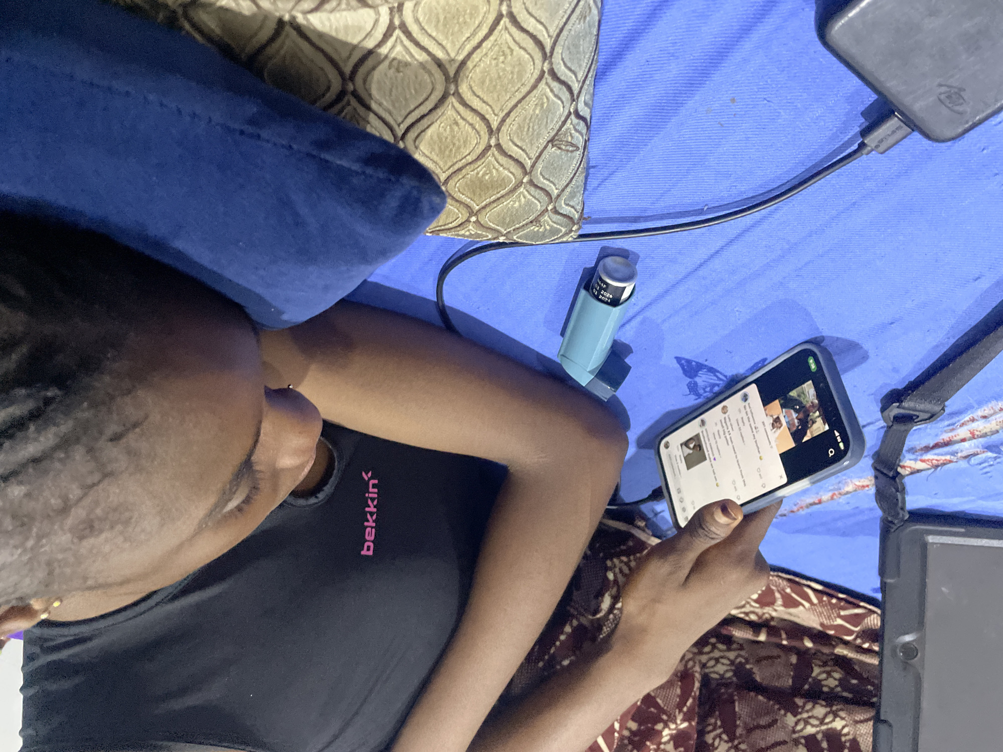 In Abuja, Nigeria, a person is lying on a blue surface, possibly a bed, with a patterned cloth behind them. They are looking at a smartphone held in their hand. A black tablet is positioned to the left of the smartphone. An asthma inhaler, light blue in color, is lying next to the smartphone. A black power bank is connected to the smartphone via a black cable. The person is wearing a black top with pink lettering that reads "bekkin". A blue pillow is visible to the right of the person.