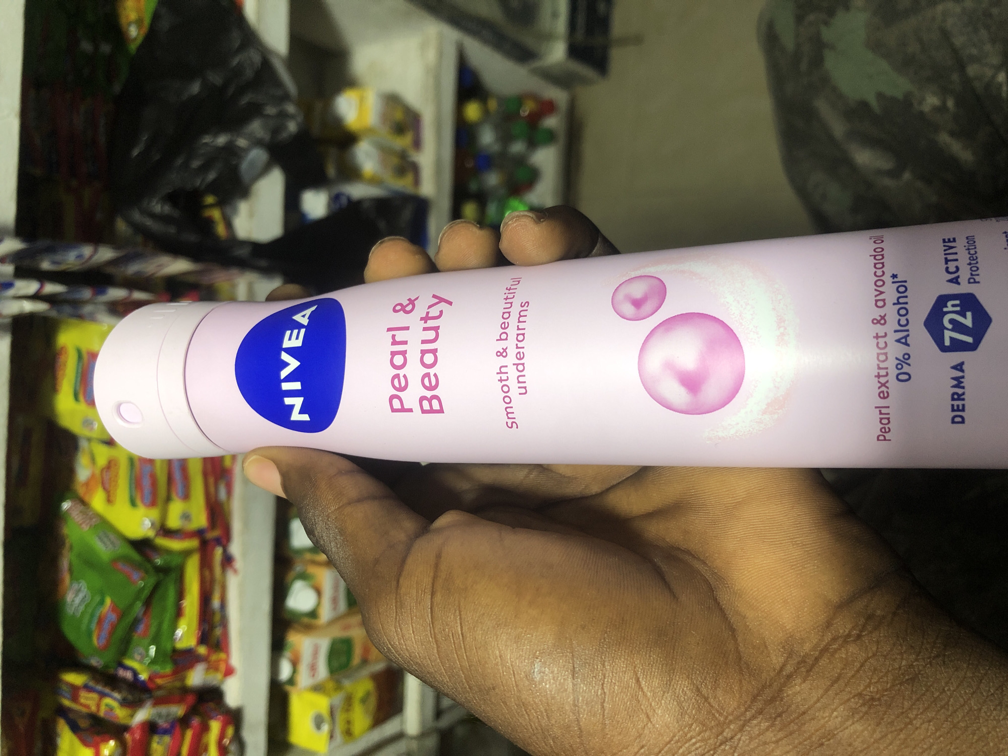 A dark-skinned hand is holding a Nivea Pearl & Beauty deodorant spray. The deodorant can is pink with blue lettering and features images of pink bubbles. Text on the can reads "Pearl & Beauty," "Smooth & beautiful underarms," "Pearl extract & avocado oil," and "0% Alcohol*." Below this, a blue hexagonal icon with "72h" and "ACTIVE Protection" is visible. The background shows shelves stocked with various products, including packaged snacks and drinks, suggesting a retail environment, possibly a small shop or kiosk in Ado, Nigeria. The lighting is somewhat dim, and the overall scene is candid and casual.