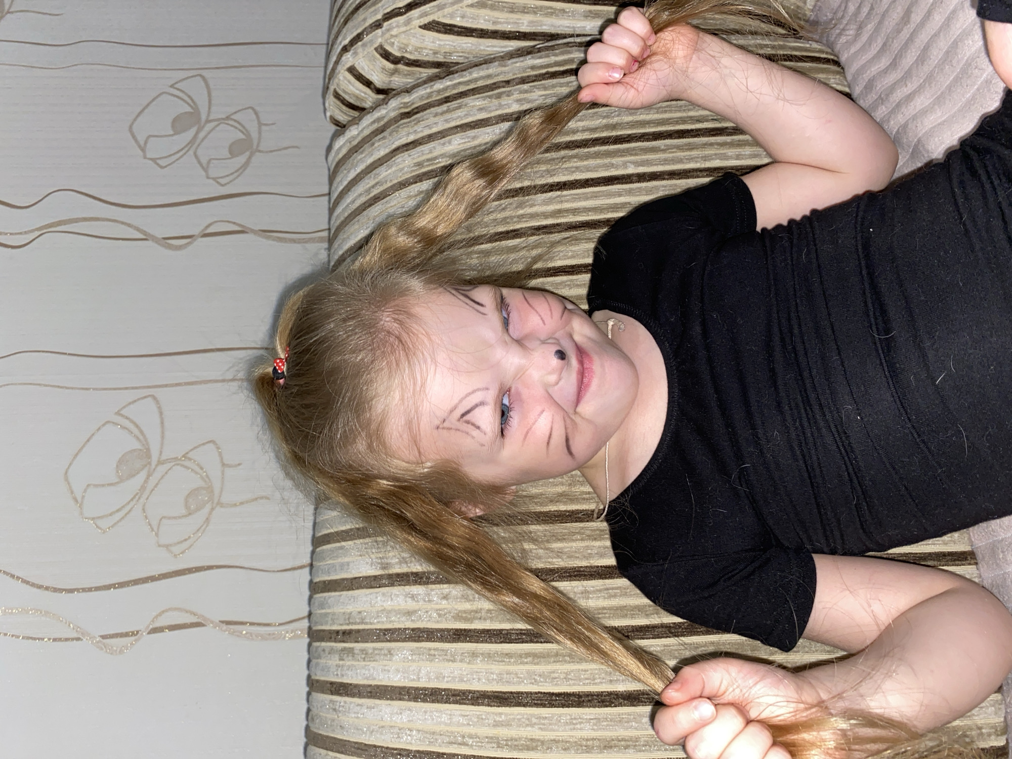 A young child with blonde hair, styled in two pigtails secured with small red and white polka dot hair ties, lies playfully on a striped cushion or couch. The child is wearing a black long-sleeved top and has black face paint applied to resemble a cat, with whiskers, a black nose tip, and pointed markings above the eyes. A wide grin spreads across the child's face, conveying a sense of fun and happiness.
The child is positioned diagonally across the frame, looking towards the viewer. Both hands are holding onto strands of her pigtails. The cushion or couch features prominent horizontal stripes in shades of brown, beige, and light grey or metallic silver. To the left, a light-colored wallpaper is visible, decorated with subtle, shimmering wavy lines and two stylized, outlined eye-like shapes. The setting appears to be indoors, likely a home in Votkinsk, Russia, under artificial lighting. There is no visible text in the image.