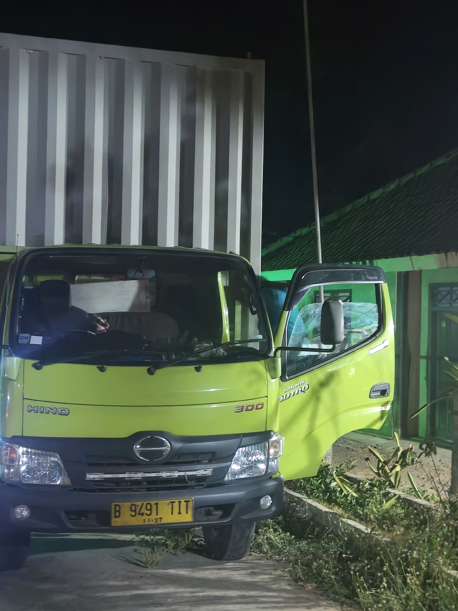 A light green Hino 300 Dutro truck is parked on a paved surface at night, partially obscured by a large, ribbed white shipping container. The truck's headlights are on. The driver's side door is open, revealing a driver seated inside wearing a dark shirt and holding a red object. The truck's license plate is visible and reads "B 9491 TIT" with "11-27" below it. In the background, a green building with a dark, sloped roof is visible, along with some overgrown vegetation. The scene is likely in a commercial or industrial area in Kroya, Indonesia, given the vehicle's registration plate format and the common use of Hino trucks in the region.