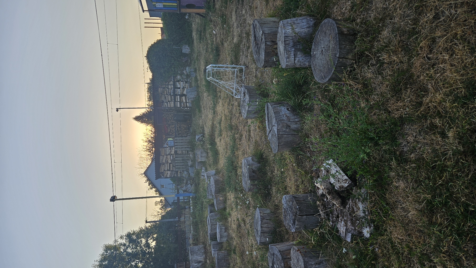 The image depicts an outdoor scene in the city of Prešov, Slovakia, with a focus on a natural playground area. In the foreground, numerous tree stumps of varying sizes are arranged in an informal pattern, serving as seating or stepping stones. The ground is covered with dry grass and some patches of green vegetation. In the midground, a small, white, foldable goal net is visible. Behind the net, a stack of firewood and a small blue and white building are situated. Further in the background, trees and more stacks of firewood are present, with a hazy sunset visible in the distance. Electrical wires and poles are also noticeable against the sky. The overall impression is one of a rustic, somewhat unkempt outdoor space.