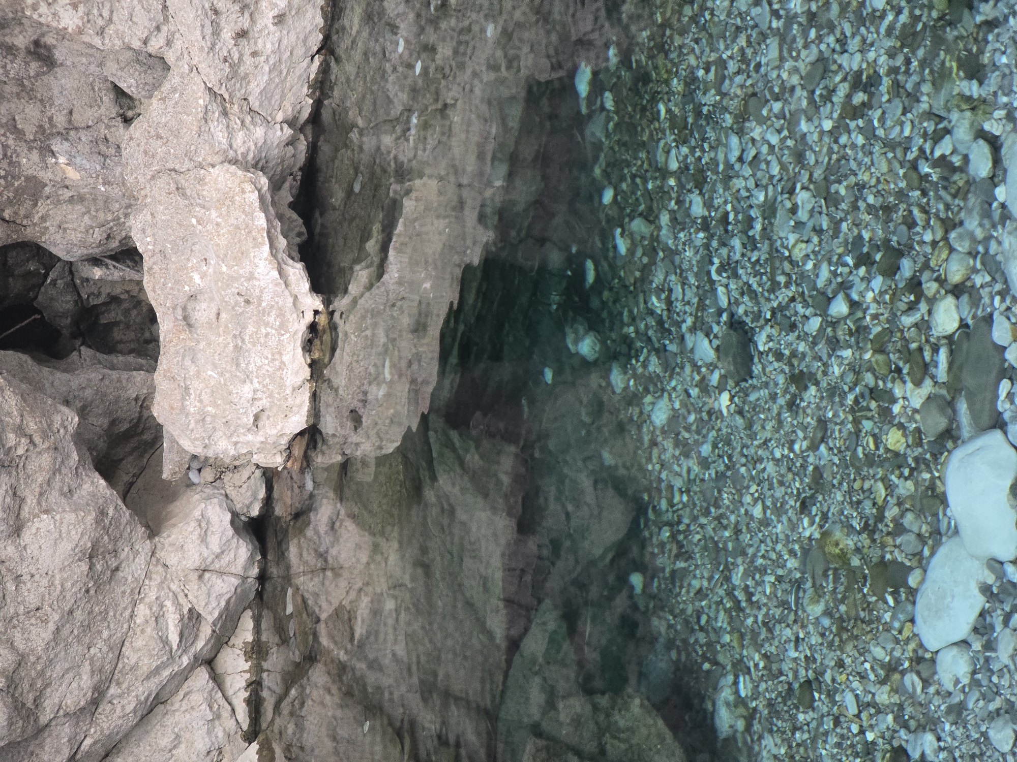 The image shows a shallow, clear body of water with a rocky bottom. The water's surface reflects the large, light-colored rocks that form the surrounding cliff face. The rocks are a mix of smooth and jagged textures, with crevices and overhangs visible. The water is a turquoise color, and the rocky bed is visible through the clear liquid. Some of the rocks on the seabed are smooth and rounded, while others are more angular. The environment appears to be a natural setting, possibly a riverbed or a small cove. The location is identified as Bioče, Montenegro. There are no people or animals visible in the image, nor any signs of human activity.