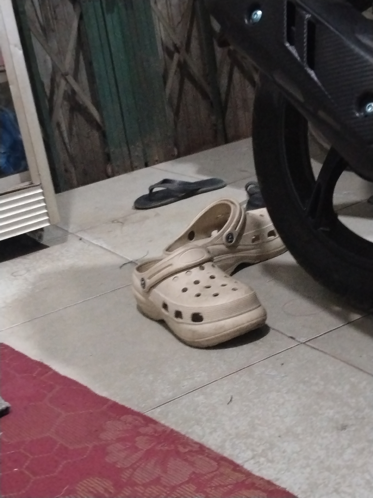 In Unknown, Indonesia, a pair of beige Crocs sits on a tiled floor next to a pair of black flip-flops. A red rug with a hexagonal pattern is partially visible in the foreground. A motorcycle's rear tire and exhaust system are to the right, with a portion of its black, textured fender visible above the tire. The background shows a closed metal gate with vertical slats, and a white-framed window with horizontal blinds on the left. The lighting suggests an indoor or covered outdoor area.