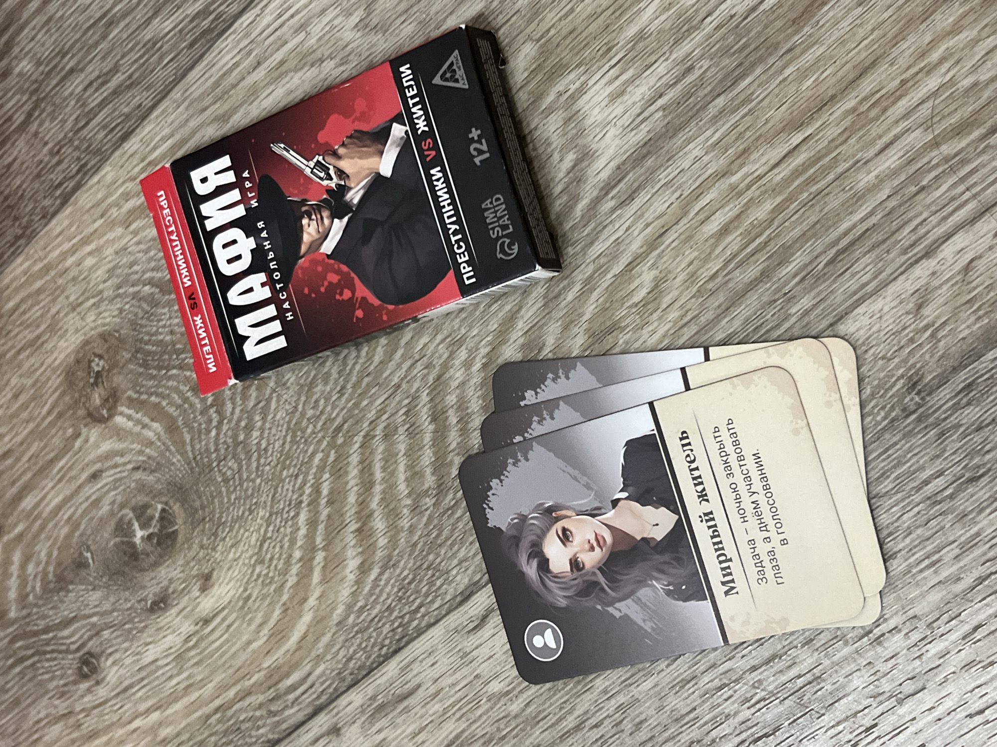 The image shows a close-up of a board game called "Mafia" on a wooden floor. The game box is red and black, featuring an illustration of a man in a suit holding a gun. The cards in the foreground depict illustrated characters, with one card clearly showing a woman with gray hair and the text "Мирный житель" (Peaceful Resident) and a description of her role in the game. The environment appears to be indoors, with the texture of the wooden floor being prominent. There is no indication of the location being in Дачное, Russia, or any other specific city. The image is a still photograph, with no people actively interacting with the game in the frame.
