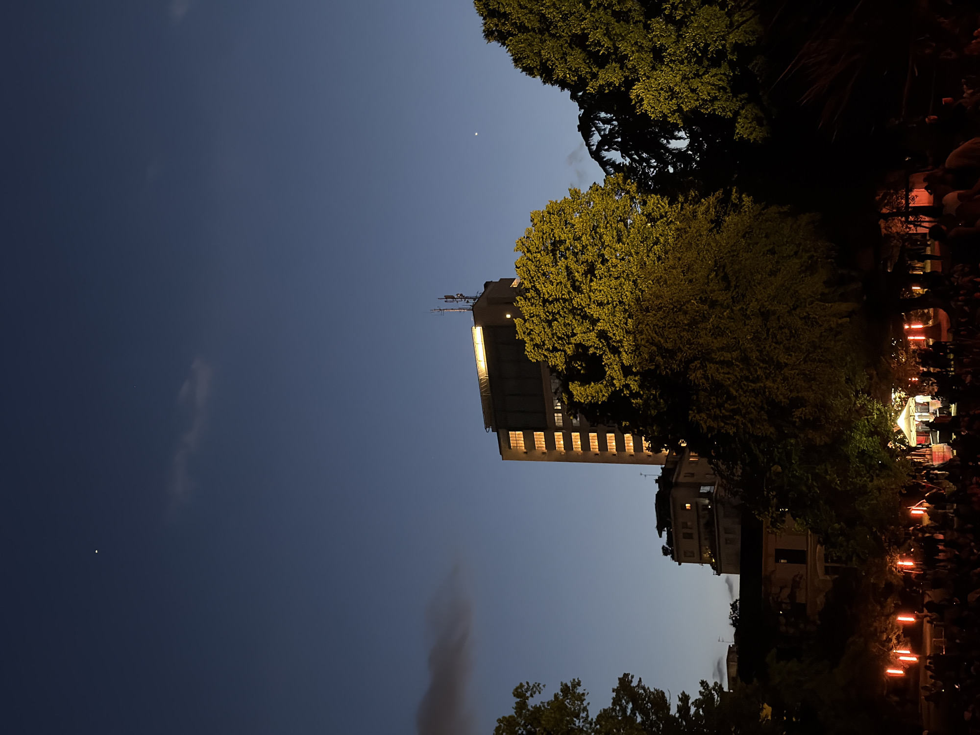 At night, in Milan, Italy, a tall building is brightly lit while nearby trees appear dark. The sky is dark blue. Two small dots, possibly stars, are visible in the sky. Below the building, another building is visible, partially obscured by trees. At ground level, there are some sources of warm light, possibly small fires. People are dimly visible near the light.
