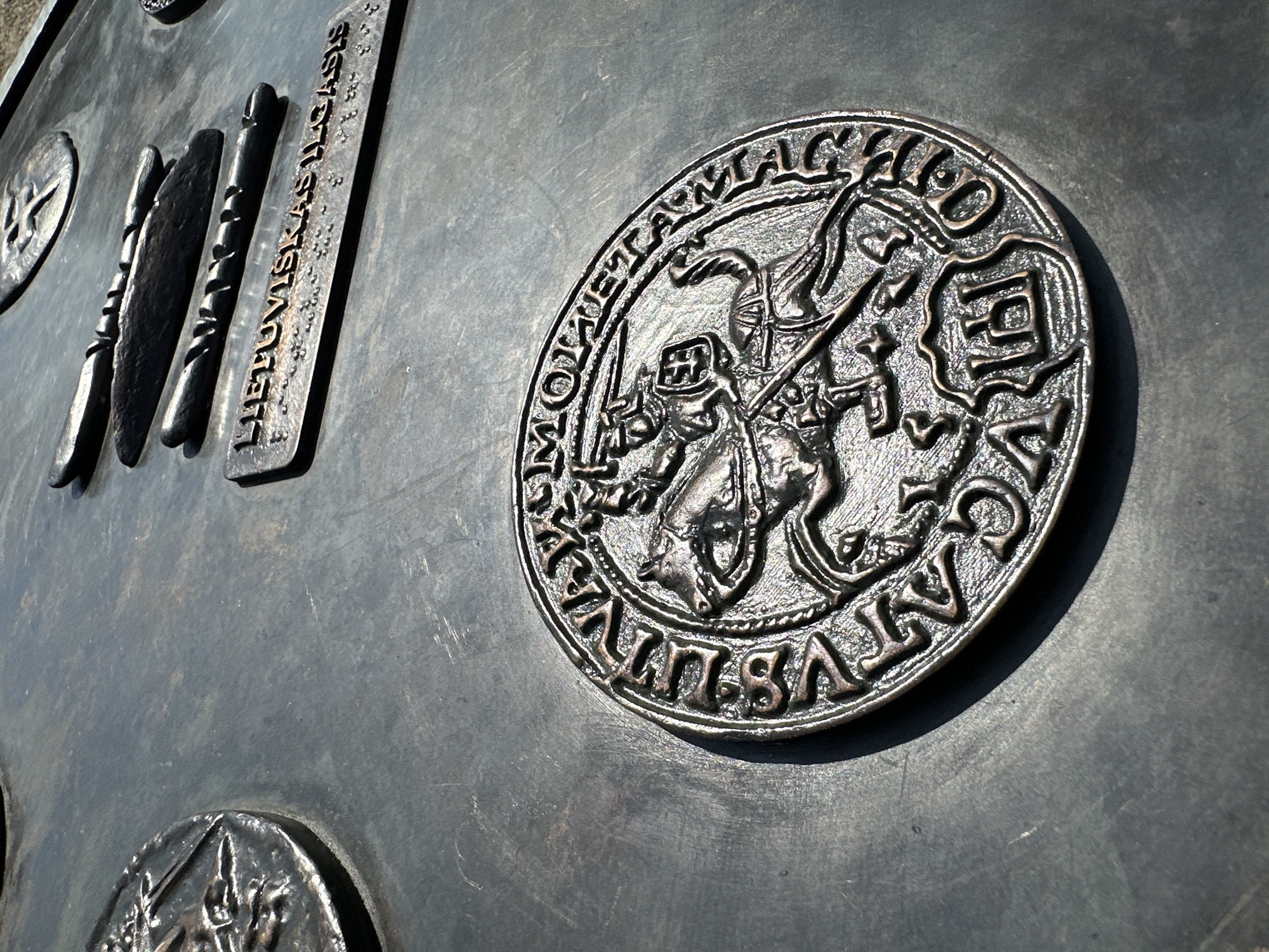 The image shows a close-up of a bronze relief on a dark, textured surface, likely part of an outdoor monument or information board. In the foreground, a circular bronze medallion features a detailed relief of a knight on horseback, a symbol commonly associated with Lithuania, specifically the "Vytis" or "Pahonia" motif. The knight is depicted with a sword raised, and a shield bearing an emblem. The medallion is surrounded by text in an archaic script, partially obscured.
In the middle ground, a rectangular bronze plaque is visible with raised text that reads "LIETUVISKAS ILGASIS" followed by what appears to be Braille. Above this plaque are three horizontal, elongated bronze elements, resembling stylized representations of traditional Lithuanian tools or weapons.
In the background, another circular bronze element with relief details is partially visible, suggesting this is part of a larger artistic installation. The lighting indicates an outdoor setting with strong sunlight casting shadows.
This monument is located in the city of Trakai, Lithuania, specifically at the Trakai Island Castle complex. The reliefs likely serve to inform visitors about Lithuanian history, culture, and symbols.