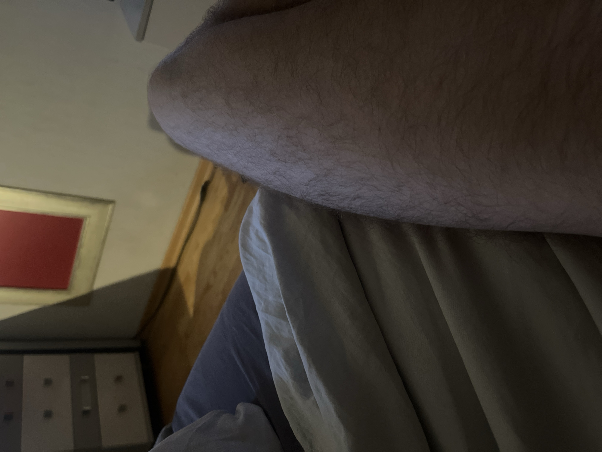 The image shows a close-up view of a person's bare leg resting on a bed with white sheets. The leg is hairy, and the skin is light-toned. The background is dimly lit and shows a dresser with drawers and a framed picture on the wall. The environment appears to be a bedroom, likely in a residential setting. The image does not provide any specific information about the city of Minsk, Belarus, or any context related to a crime scene.