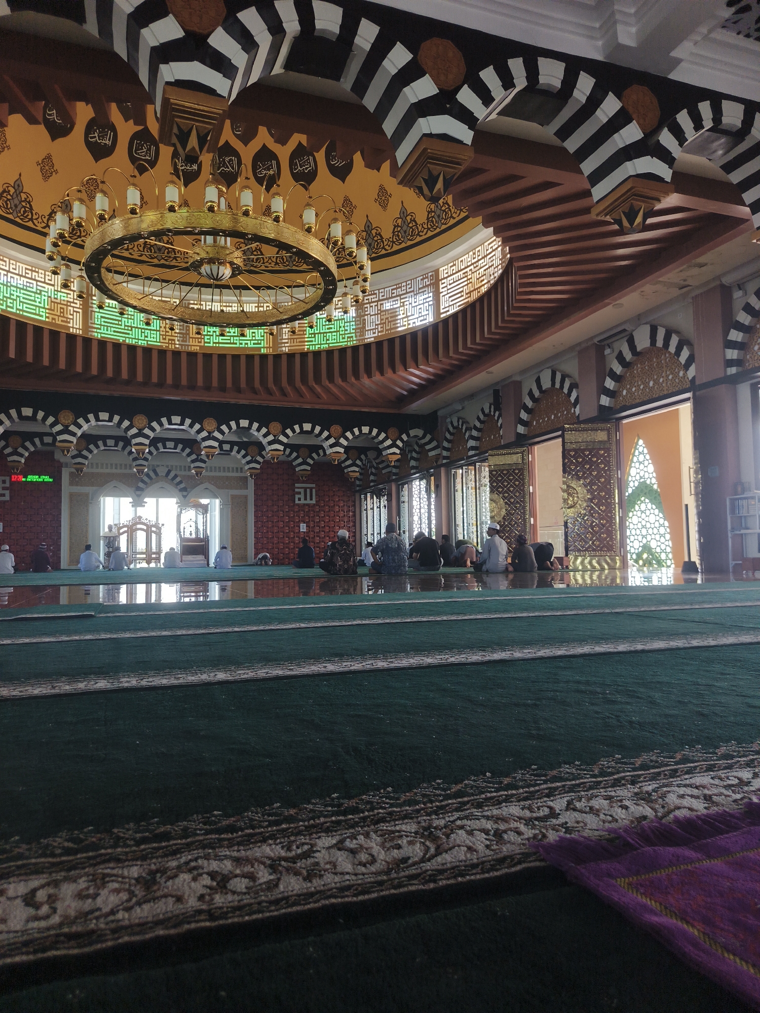 The image depicts the interior of a mosque, specifically the prayer hall, in Beringin, Indonesia. The perspective is low, angled from the green prayer carpet which is marked with white lines. In the foreground, a portion of a purple prayer mat is visible. The main area of the hall features multiple rows of people, mostly men, seated or prostrating in prayer. The architecture is ornate, with a large, circular, golden chandelier hanging from the domed ceiling. The ceiling is decorated with black and white striped arches and calligraphy. Sunlight streams in from arched doorways at the far end of the hall, illuminating patterned screens. There are also decorative tiled walls and large wooden doors with intricate metalwork. A digital clock displaying "12:21" is visible on the wall to the left. Bookshelves are present on the far right. The overall atmosphere suggests a serene and communal prayer service.