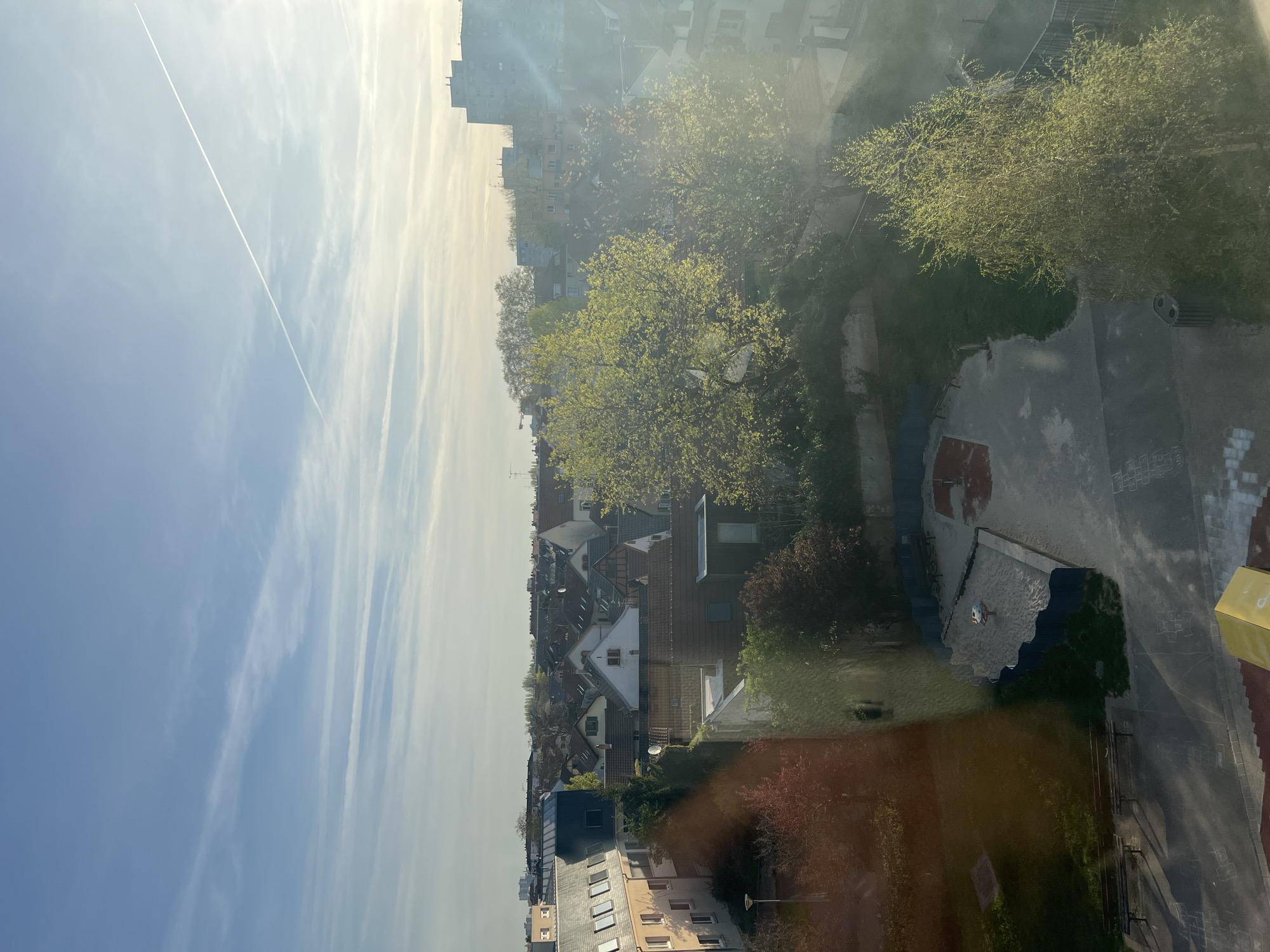 The image, taken from a high vantage point, depicts a cityscape. The sky is blue with contrails from airplanes and bands of wispy clouds. Below, residential buildings are interspersed with trees in varying shades of green. A playground with a sandbox and a paved area with painted games is visible. The location is indicated to be Schiltigheim, France. No people are clearly visible.