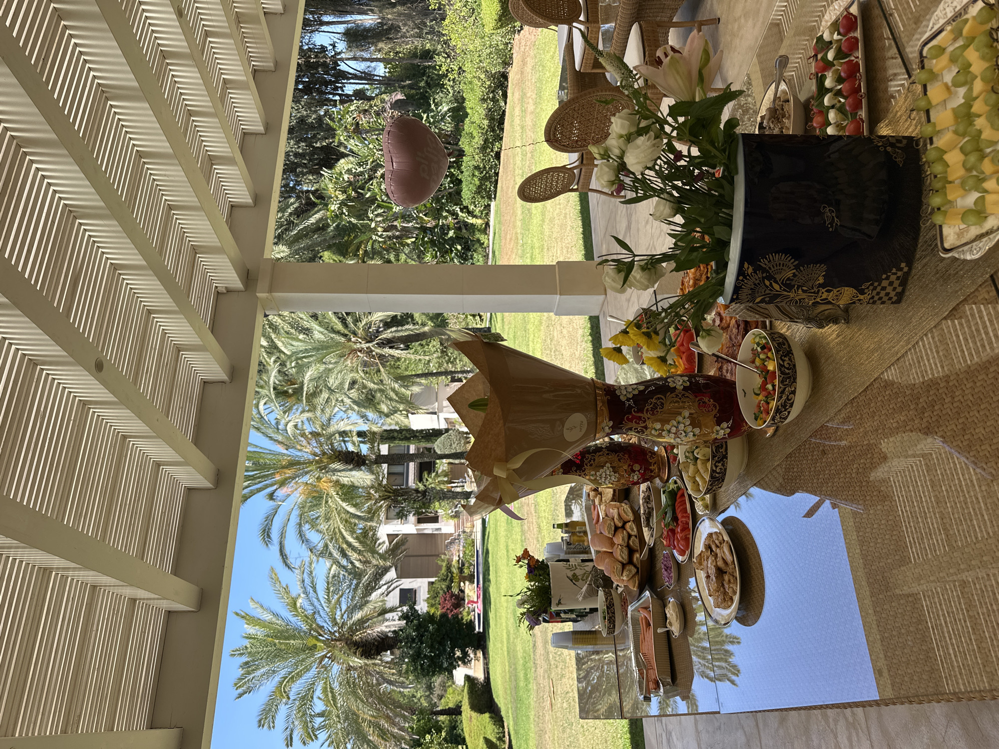 The image depicts a lavish outdoor dining spread set on a glass-topped table on a patio. The table is laden with various food items, including platters of sausages, bread rolls, salads, fruit skewers, and other dishes. Decorative vases and floral arrangements add to the festive atmosphere. In the background, a lush green lawn is visible, dotted with palm trees and a swimming pool, suggesting a residential property in an affluent area. The architectural style of the house in the background, with its modern design and large windows, is consistent with properties found in Savyon, Israel, known for its upscale residences and spacious gardens. A heart-shaped balloon floats gently in the air, adding a touch of celebration to the scene. The overall impression is one of a well-appointed outdoor gathering at a private residence in Savyon.