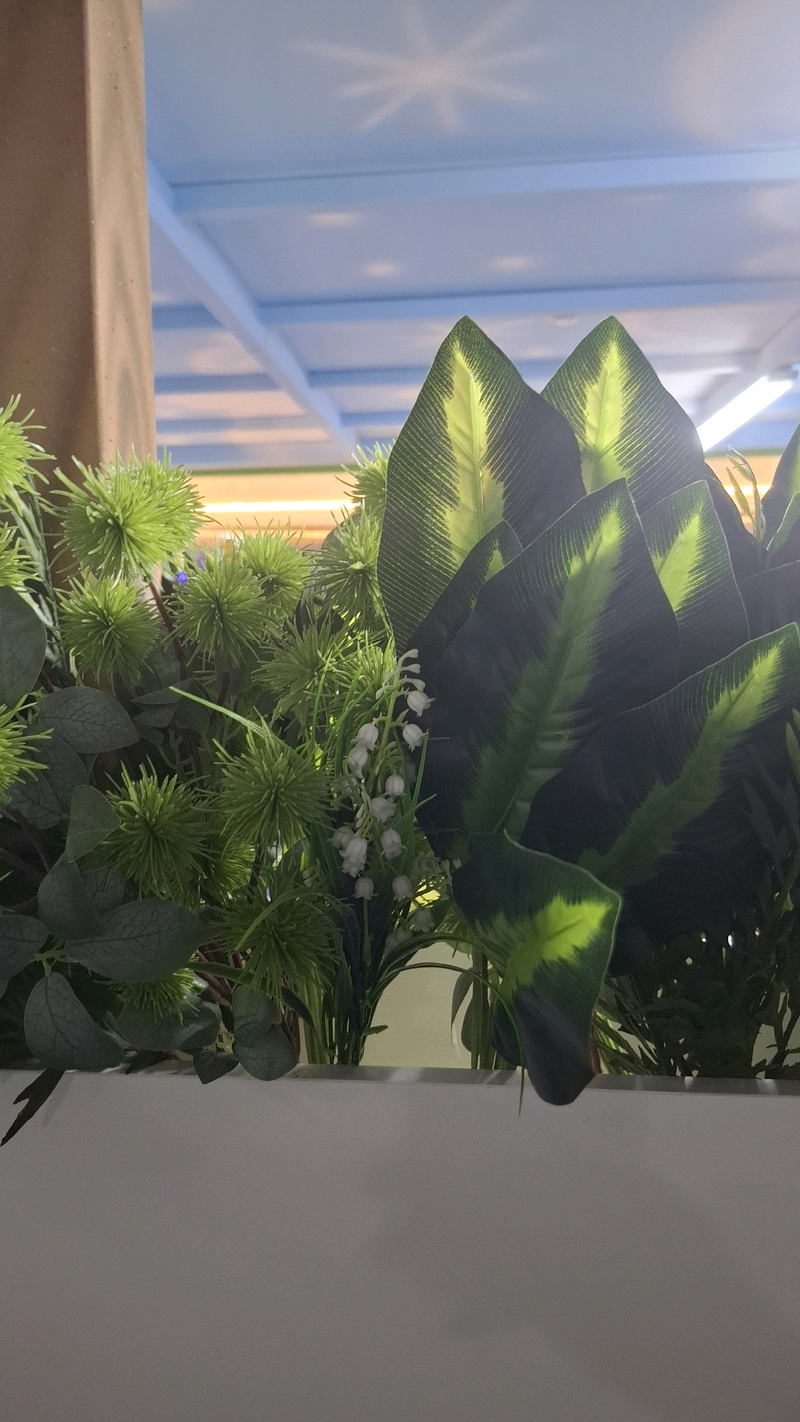 The image shows an artificial plant arrangement in a white planter. The arrangement includes large, dark green leaves with yellow-green stripes, spiky green floral accents, and small white flowers resembling lily of the valley. In the background, a light blue ceiling with a geometric pattern is visible, and a light-colored column is to the left. The setting appears to be indoors, possibly a commercial space or a home in Shymkent, Kazakhstan, though there are no specific landmarks to confirm the exact location within the city. There are no people or actions depicted.