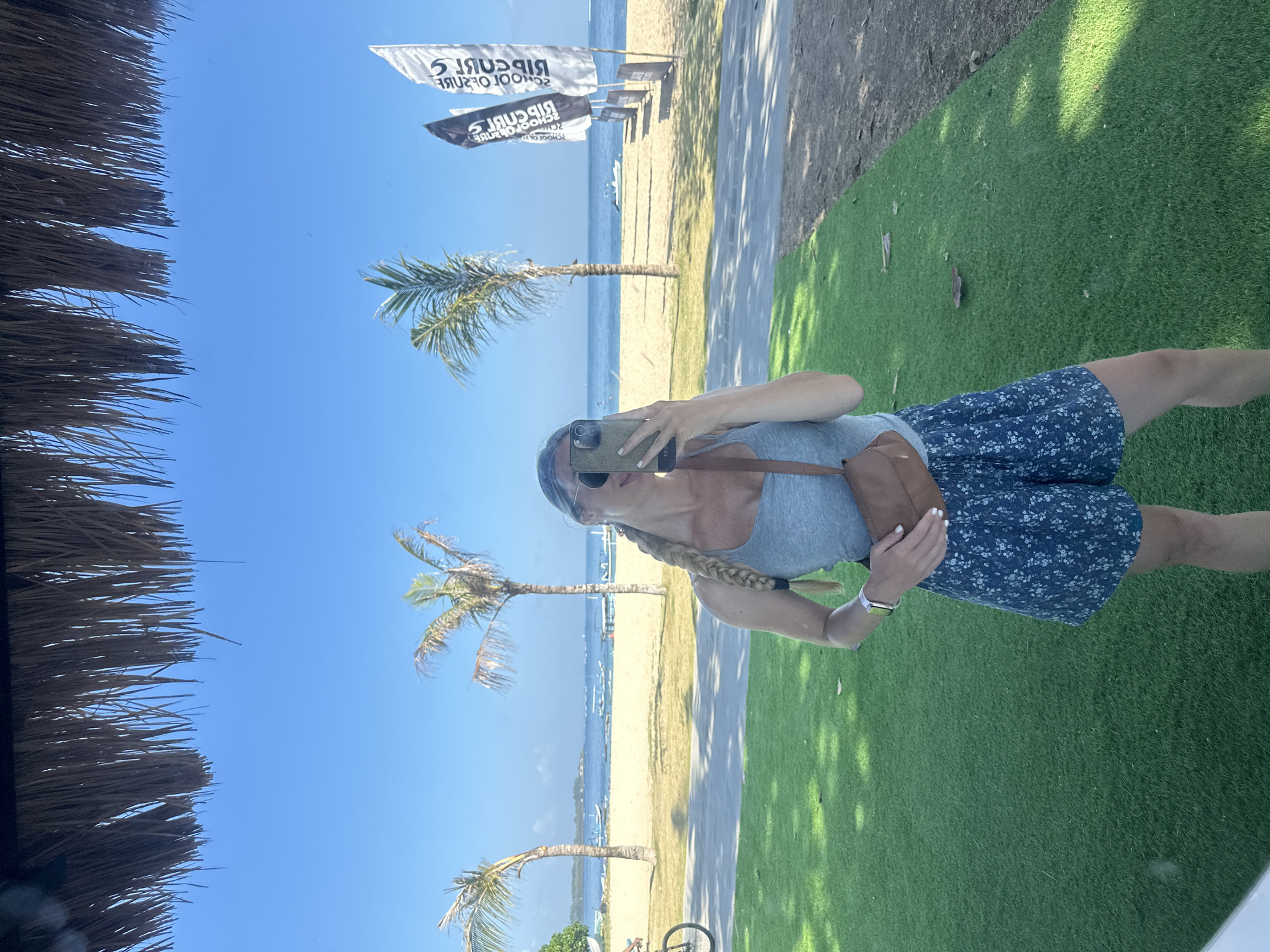 A woman with blonde hair in a braid is taking a selfie in a mirror. She is wearing a gray tank top, blue floral shorts, sunglasses, and a brown fanny pack across her chest. She is standing on artificial green grass, with a sandy beach, palm trees, and the ocean in the background. Three flags are visible on the beach. The scene is outdoors on a clear, sunny day. The location is a beach in Denpasar, Indonesia.