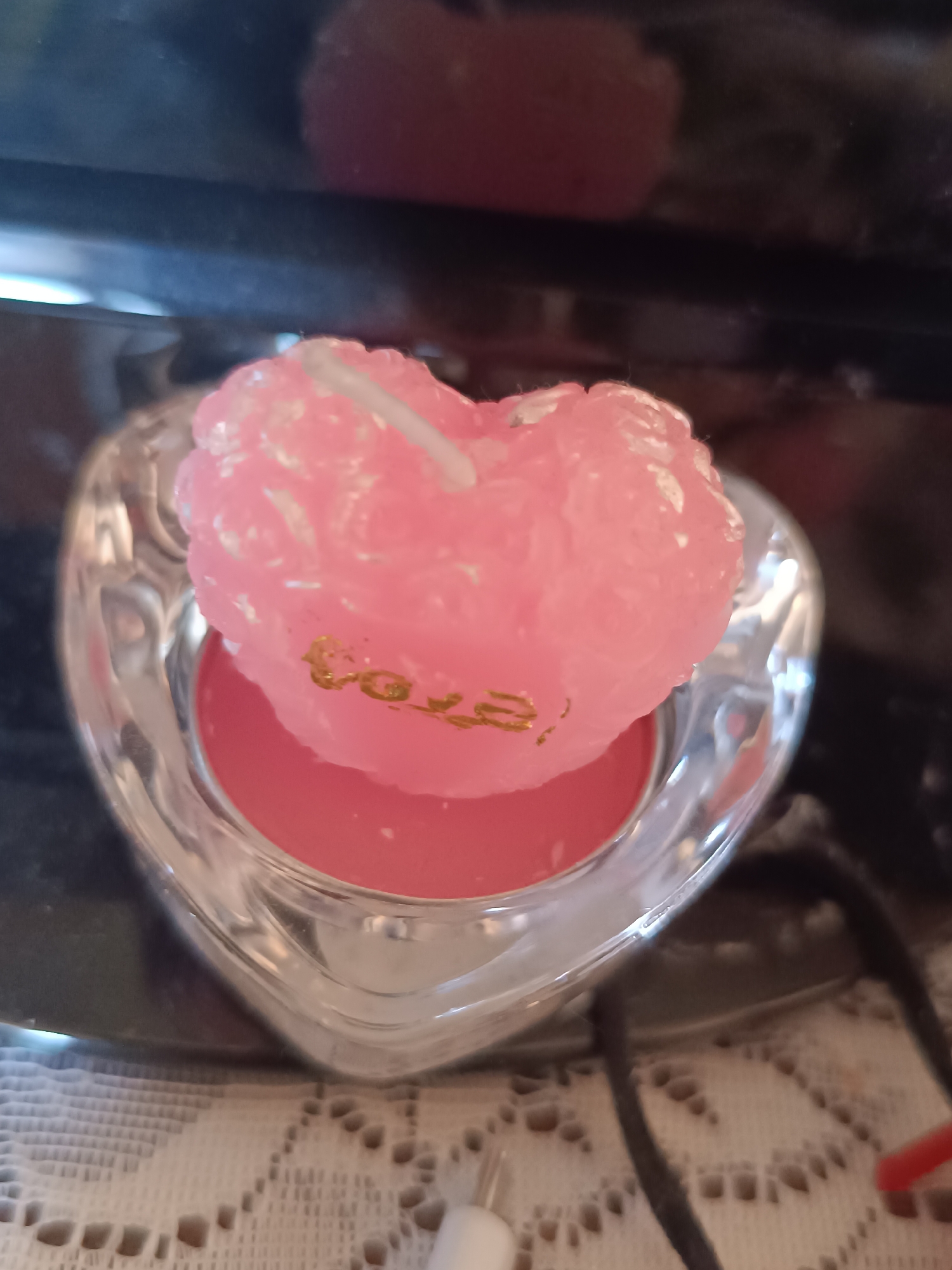 A close-up shot shows a pink, heart-shaped candle resting inside a clear, heart-shaped holder. The candle has the word "love" written on it in gold lettering. The holder appears to be sitting on a surface covered with a white, lacy tablecloth. Black and red wires are visible in the lower right corner of the image. The location is Unknown, Unknown.