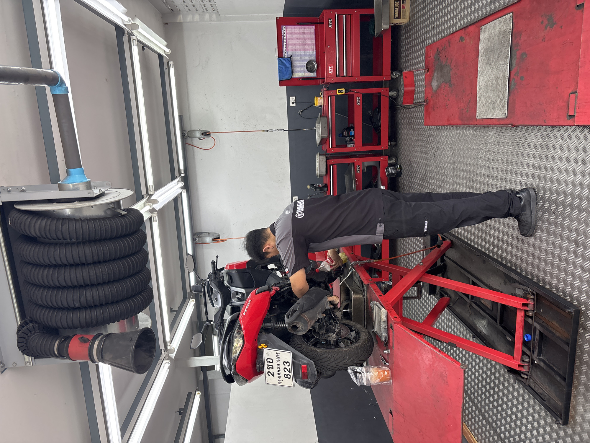 The image depicts an indoor auto repair shop. A man wearing a dark gray Yamaha shirt and black pants is working on a red motorcycle positioned on a red motorcycle lift. The bike has a license plate displaying "2 ขอ กรุงเทพมหานคร 823." Above the motorcycle, there's a large black exhaust extraction system with a black funnel-shaped end.
Behind the mechanic, there are several red tool chests with the brand name "KTC" visible. A metal diamond-patterned floor covers the shop. Fluorescent lights are mounted on the walls and ceiling.
Based on the license plate, the motorcycle is registered in Bangkok, Thailand. While the photo does not provide location information in the city of Baan Bo Phut, Thailand, the license plate may mean this photo was taken somewhere else.