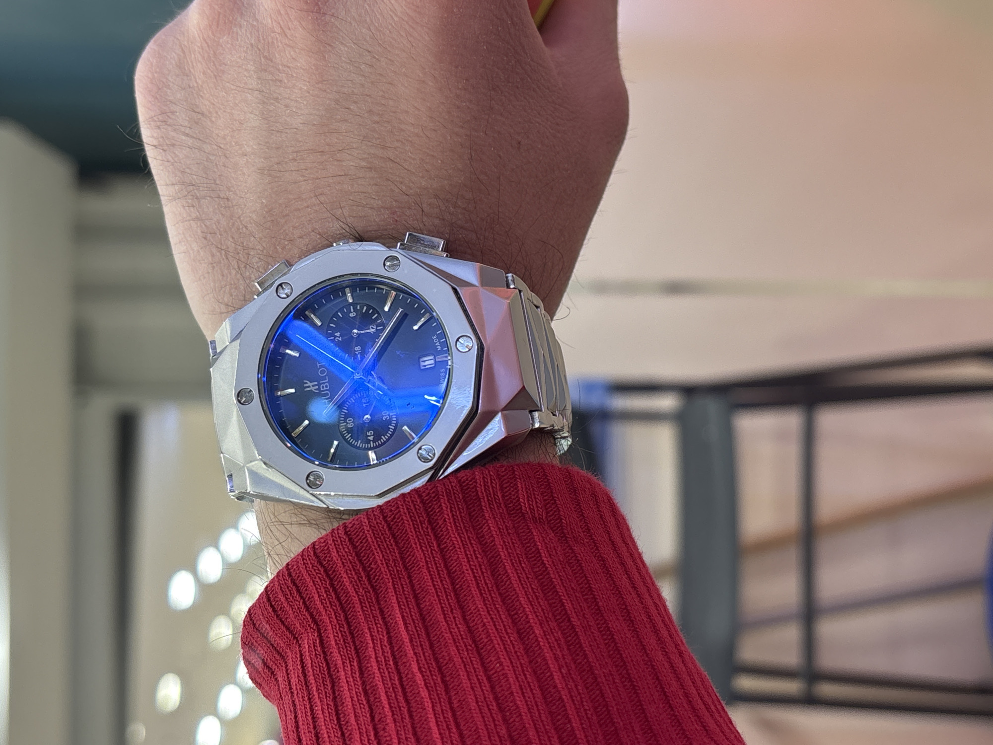 A close-up image shows a wrist wearing a silver Hublot watch with a blue face. The watch is prominently featured, with a red, ribbed sleeve partially covering the wrist. The background is blurred, showing hints of an interior space with lights and possibly a window or display. The location in Saint Petersburg, Russia is not discernible from the image.