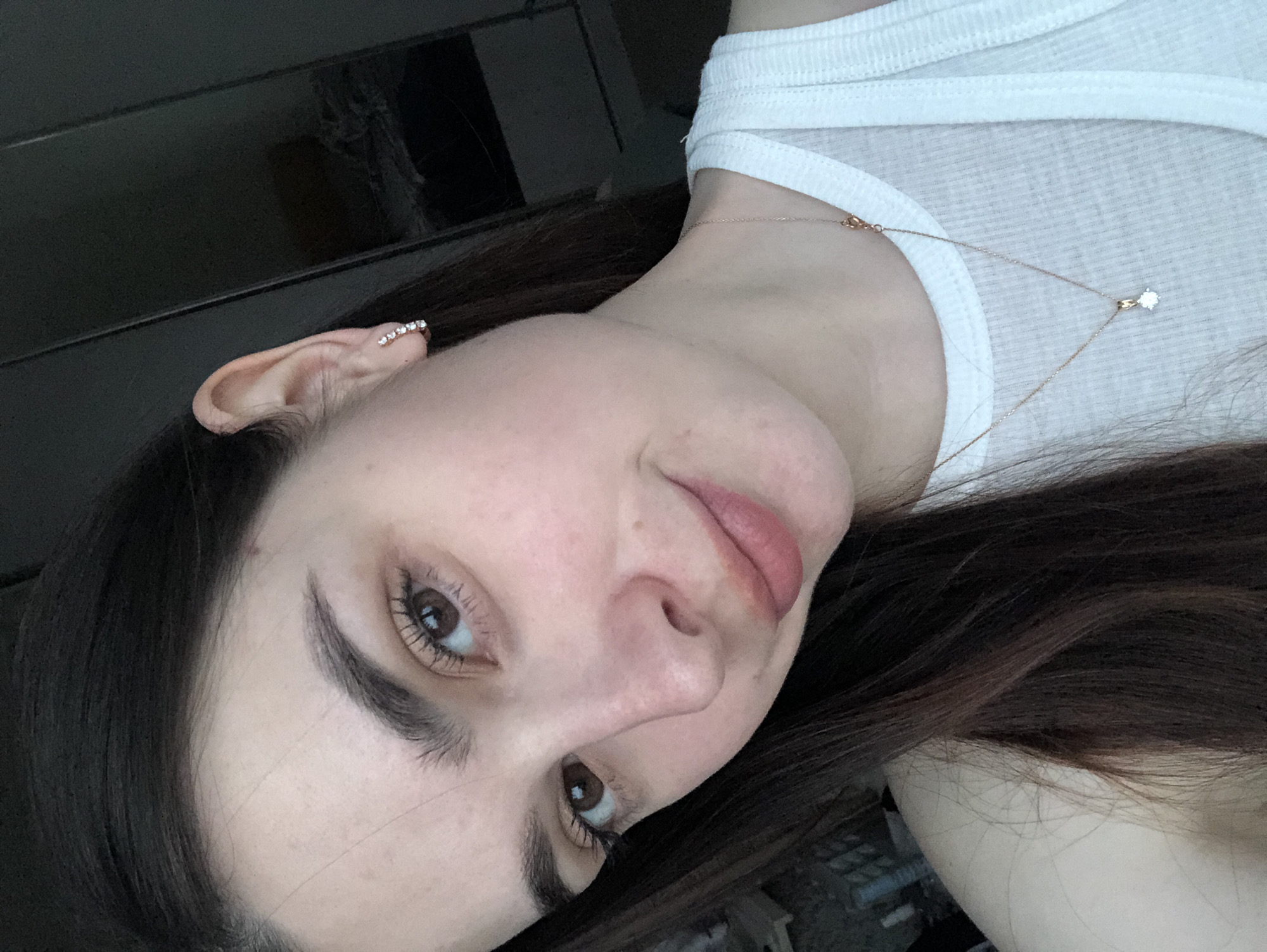 A close-up, slightly angled selfie shows a woman with fair skin and brown hair. She is wearing a white tank top and a delicate gold necklace with a small diamond pendant. Her left ear has a bar-shaped earring with several small stones. She has brown eyes and minimal makeup. The background is dark and out of focus, with a hint of a window frame visible in the upper left corner and what appears to be a patterned floor in the lower right. No information indicates this photo was taken in Noviy, Russia.