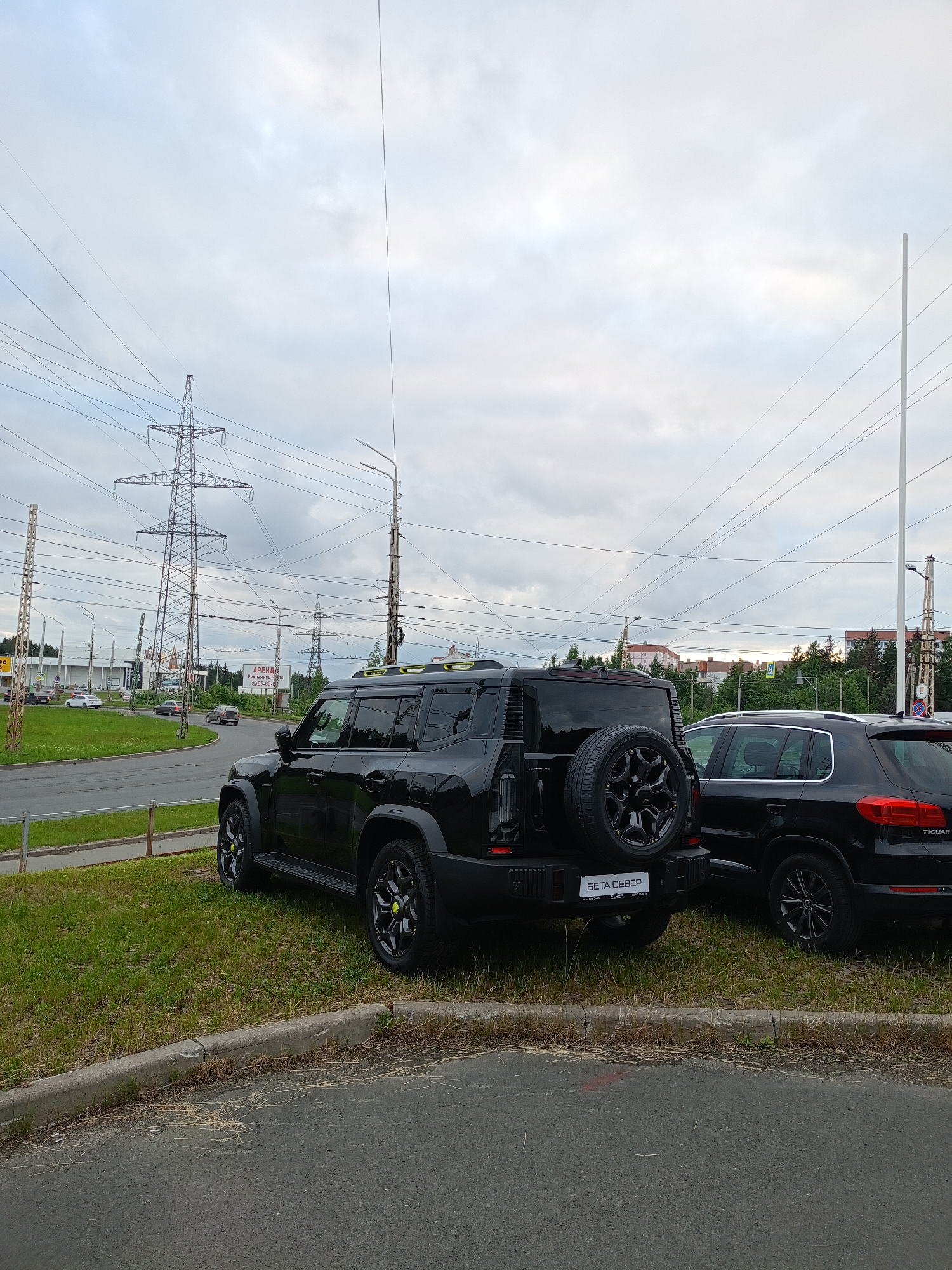 Two black SUVs are parked on a grassy median next to a paved road. The SUV in the foreground is a modern, boxy vehicle with a spare tire mounted on the rear. The SUV in the background appears to be a Volkswagen Tiguan. The scene is set against a cloudy sky with tall power line towers and electrical wires visible. In the distance, there are buildings and trees, indicating an urban or suburban environment. A sign in Russian with the word "АРЕНДА" (ARENDA), meaning "rent," is visible on a billboard in the middle ground. The location appears to be a commercial area with a road and a grassy strip. Based on the presence of the sign in Russian and the general appearance of the landscape and infrastructure, the scene is likely in Petrozavodsk, Russia. No people are visible in the image.