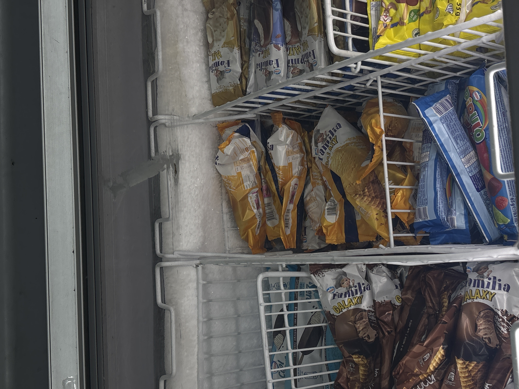 This is a photograph taken inside a freezer, likely in a retail establishment. The freezer is stocked with various ice cream products. The shelves are white wire racks. The left side of the freezer is heavily frosted with ice.
Several different brands and types of ice cream are visible, including "Familia" brand ice cream in "GALAXY" and other varieties. These ice cream products are packaged in colorful wrappers and cones. Some wrappers display nutritional information and barcodes. Other visible brands include "Pirulo" and ice cream featuring cartoon characters.
The location in the city of Plovdiv, Bulgaria, cannot be determined from the image.