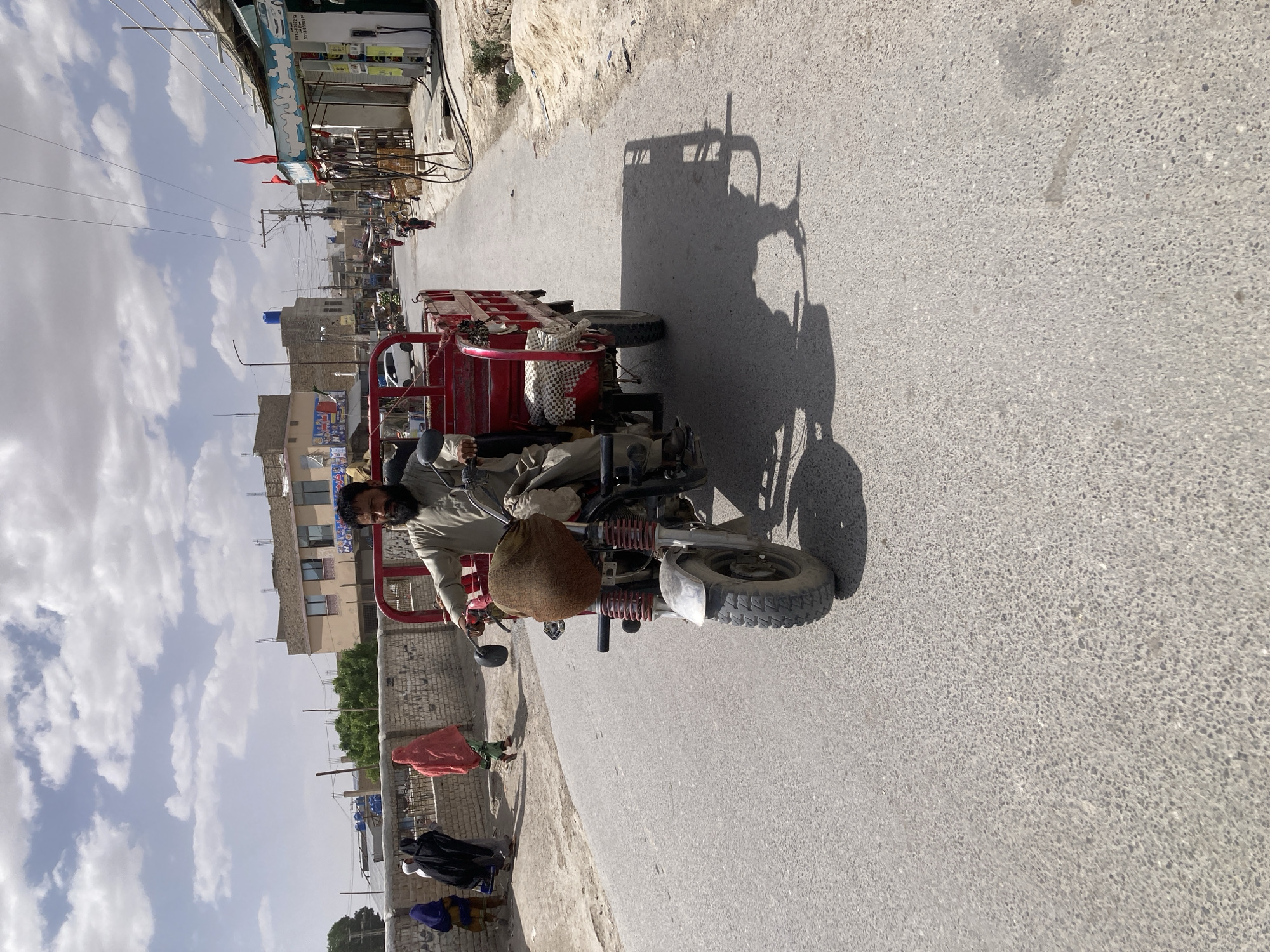 The image presents a street scene in Quetta City Tehsil, Pakistan, under a partly cloudy sky during daylight hours, likely late morning or early afternoon given the prominent shadows. The main subject is a man with a dark beard, wearing a light-colored shalwar kameez, operating a red motorized tricycle, possibly a cargo auto-rickshaw. A brown sack is visible on the front of his vehicle.
The road is asphalt, and the tricycle casts a large shadow to its right. Buildings line the left side of the street, some appearing commercial with signage and red flags. One blue and white sign is clearly visible, displaying phone numbers: "03135509748," "03003055899," "03138805375," and "03337956417," with other text partially obscured but possibly related to a "GAS POINT." Power lines and poles traverse the scene overhead. Further down the street, a blue water tank is visible on a building's roof.
On the left side of the road, several people are visible in the background, including a woman in a red garment and another in dark attire, suggesting daily foot traffic. The right side of the road appears to transition into a dusty, unpaved area. The architecture and vehicle type are consistent with the region, depicting a typical urban or semi-urban environment.