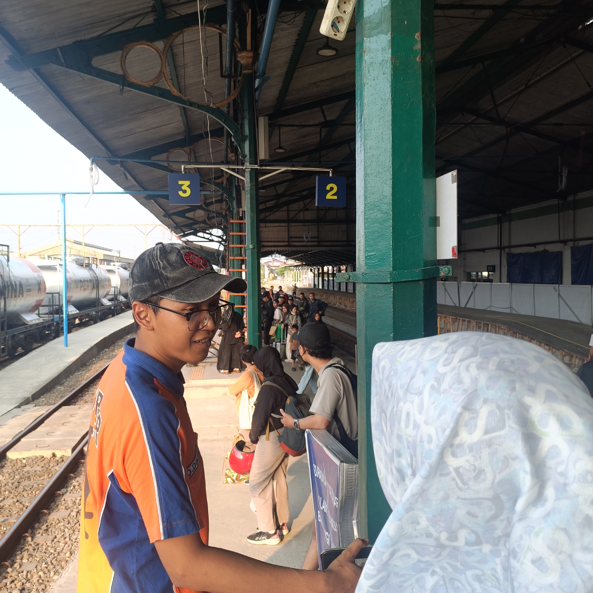 At a train station in Madiun, Indonesia, a man in an orange and blue uniform stands by train tracks, looking towards the camera. He wears a black baseball cap and glasses. Behind him, a group of people wait on a platform, with train cars visible in the background. The station has a covered roof supported by green pillars, and signs with numbers "2" and "3" are posted. The scene appears to be during daylight.