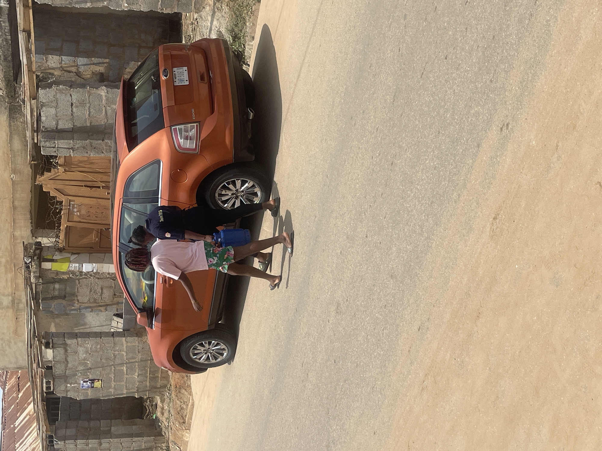 In Abuja, Nigeria, two individuals are observed walking near a parked orange Ford Edge SUV. One person, wearing a pink t-shirt and floral shorts, carries a blue container. The other individual is dressed in a black top. The scene takes place on a paved road with a dirt shoulder, adjacent to a building constructed with concrete blocks and corrugated metal roofing. The environment suggests a semi-urban or developing area.