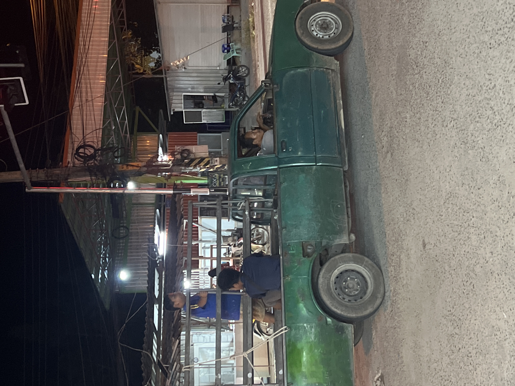 In Ko Pha-ngan District, Thailand, at night, a green pickup truck is parked on a gravel road. A man is visible in the driver's seat, holding the steering wheel. In the bed of the truck, two individuals are seated. One is wearing a blue t-shirt and shorts, and the other is wearing a dark shirt. The truck bed is enclosed by a metal frame. In the background, several motorcycles and chairs are visible near a building with a corrugated metal roof. Overhead, a complex network of electrical wires is strung between utility poles.