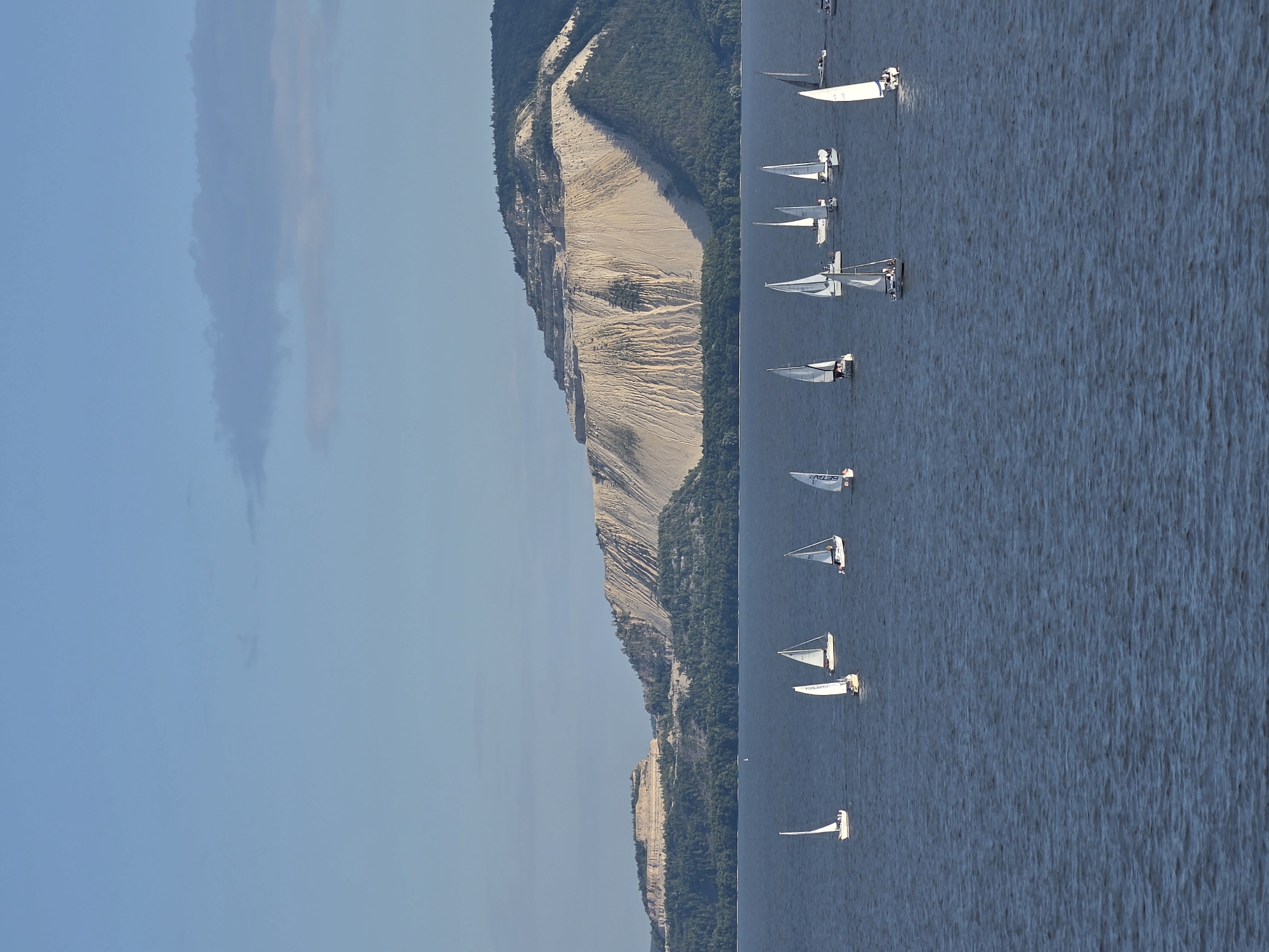 In Tolyatti, Russia, a fleet of sailboats navigates a wide, calm body of water under a clear blue sky with sparse clouds. The foreground is dominated by the deep blue water, rippled by a gentle breeze. In the middle ground, numerous sailboats, varying in size and sail color, are dispersed across the water. Some sails are white, while others are a light beige. A few individuals can be seen on the decks of the sailboats. In the background, a vast, layered sand quarry looms, its light-colored slopes scarred by excavation. This quarry is situated on a heavily wooded hill, and further in the distance, a more rounded, tree-covered hill is visible. The overall scene depicts a recreational activity taking place against a backdrop of industrial landscape.