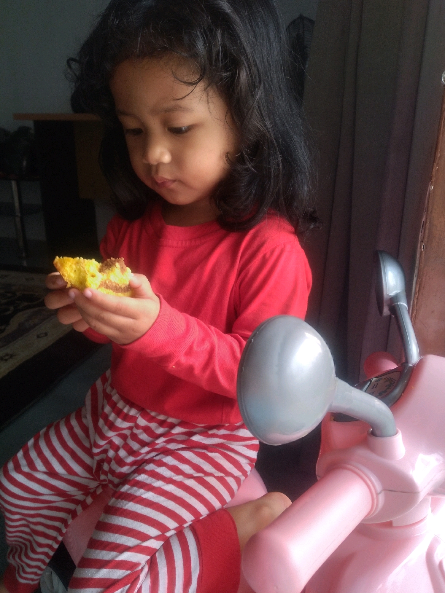 A young girl with dark, curly hair is seated and holding a piece of yellow cake with brown swirls in her hands. She is wearing a long-sleeved red shirt and red and white striped pants. She appears to be looking down at the cake. In the foreground, there is a pink toy scooter with a grey handlebar and mirrors. The background is dimly lit and shows a curtain and what appears to be furniture. The image was taken indoors, likely in a residential setting in Tembilahan Hulu, Indonesia.