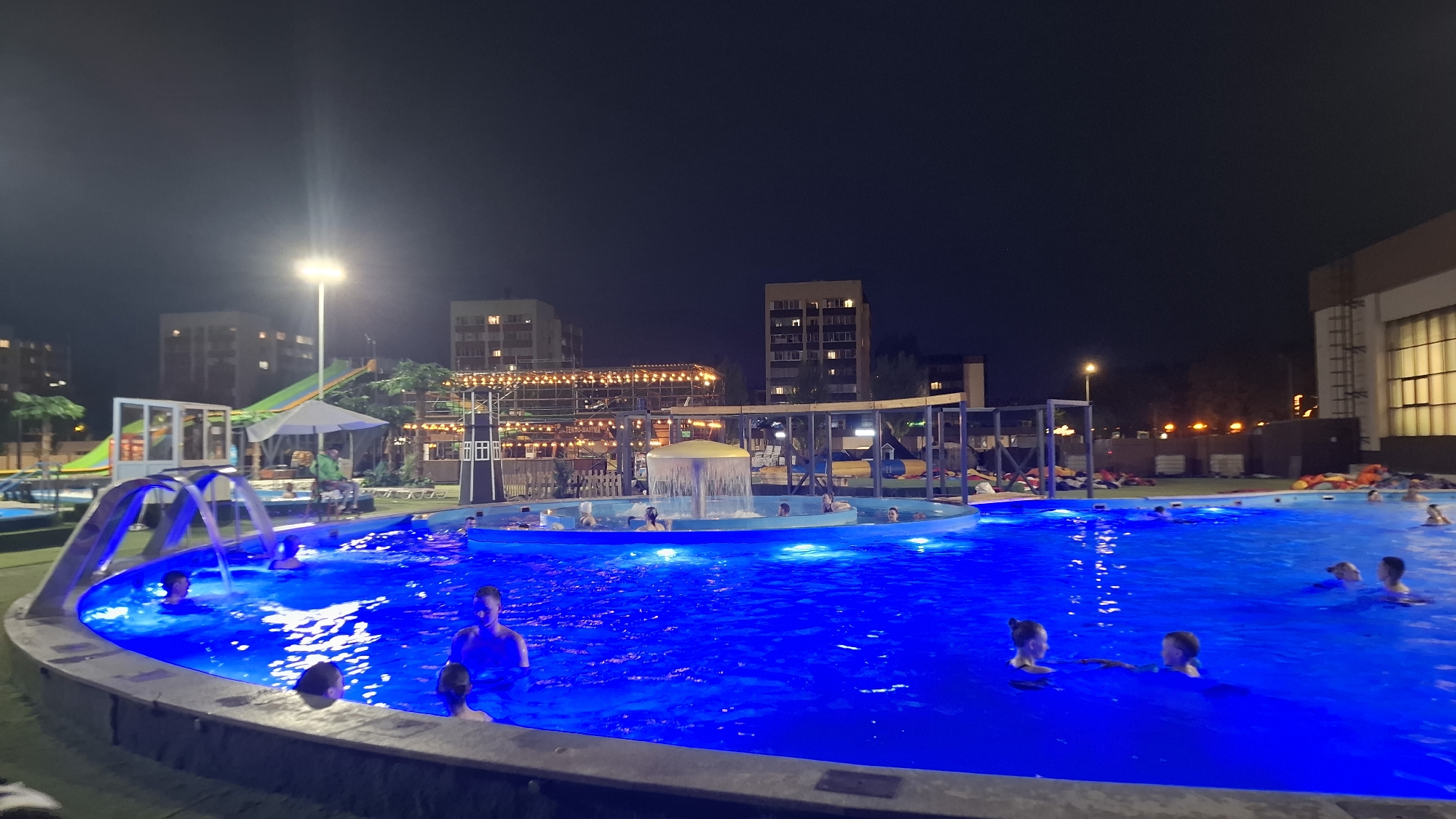 The image shows an outdoor swimming pool at night, illuminated by blue underwater lights. People are swimming and relaxing in the pool. Several architectural structures and buildings are visible in the background. The location is likely Nizhnekamsk, Russia, based on contextual information not visible in the image itself. A tall light pole is visible on the left side of the image. A water slide is located behind the pool. People are laying down on lounge chairs in the background.