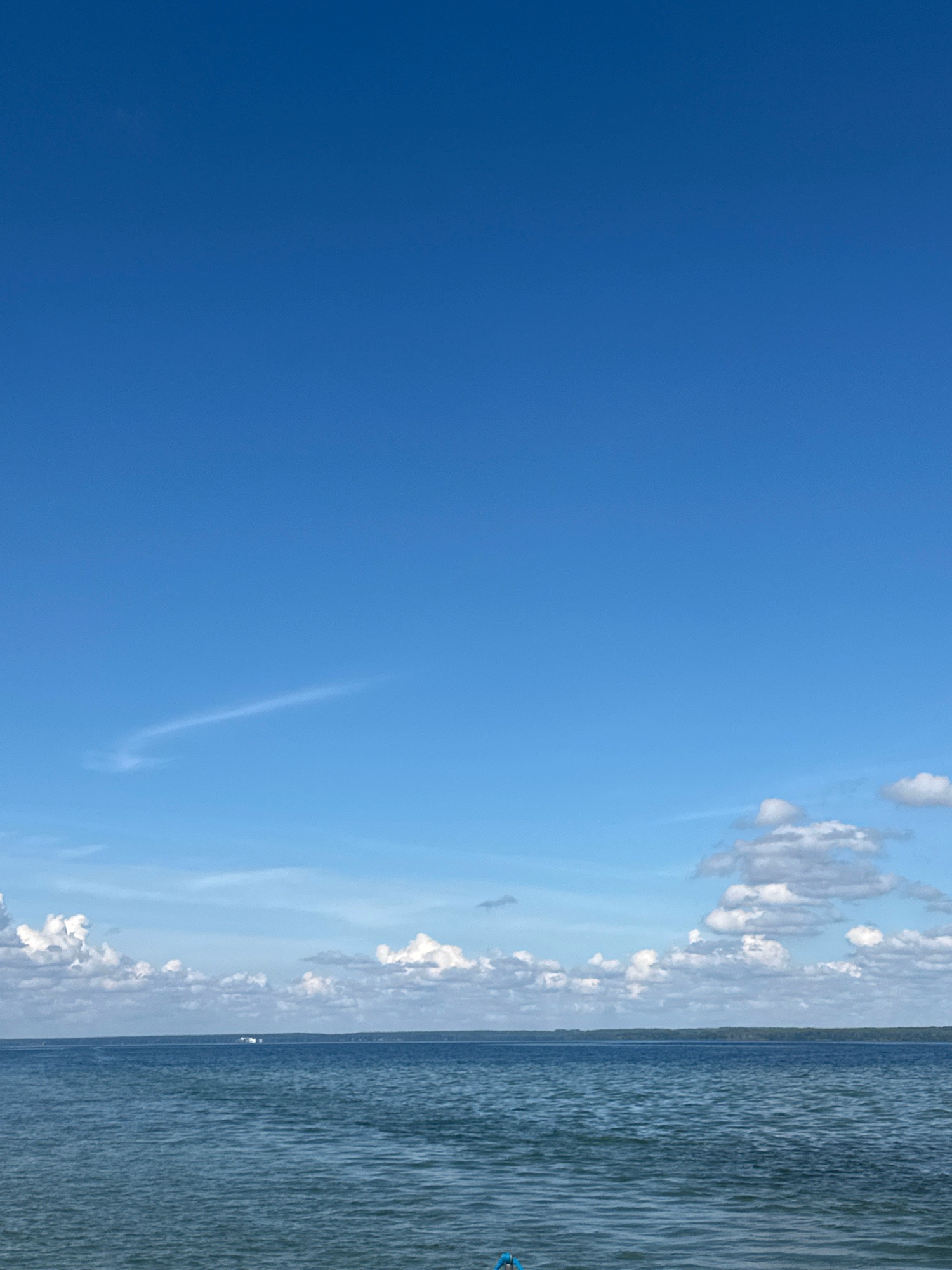 The image shows a wide, calm body of water under a clear blue sky with scattered white clouds. The water has gentle ripples. In the distance, a thin line of trees marks the horizon, suggesting a wooded shoreline. There appear to be small white objects, possibly boats or structures, on the water near the horizon line. The overall scene is serene and natural, typical of a lake or large river. The specific location is identified as the Мядзельскі сельскі Савет in Belarus.