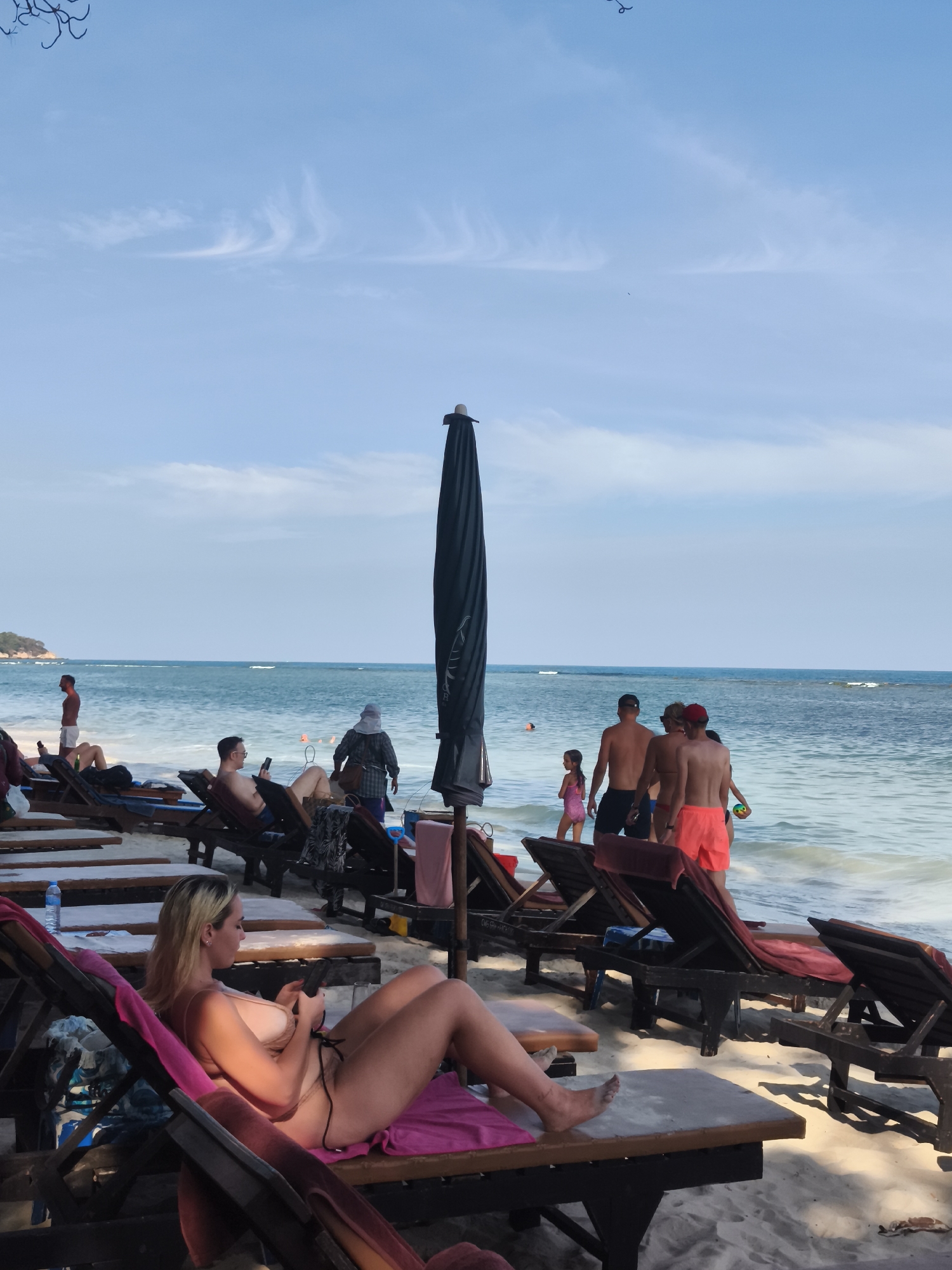 The image depicts a beach scene in the Ko Samui District, Thailand, with the ocean stretching to the horizon under a bright, partly cloudy sky. In the foreground, several empty lounge chairs are arranged on the sandy beach, indicating a typical tourist setting. A woman in a bikini is lying on one of the lounge chairs, actively using her mobile phone. Further back, a group of people are standing near the water's edge, with some appearing to be walking into the sea. Other individuals are visible reclining on lounge chairs in the midground. A large, dark beach umbrella is positioned prominently in the frame, casting a shadow. The overall atmosphere suggests a relaxed day at the beach.
