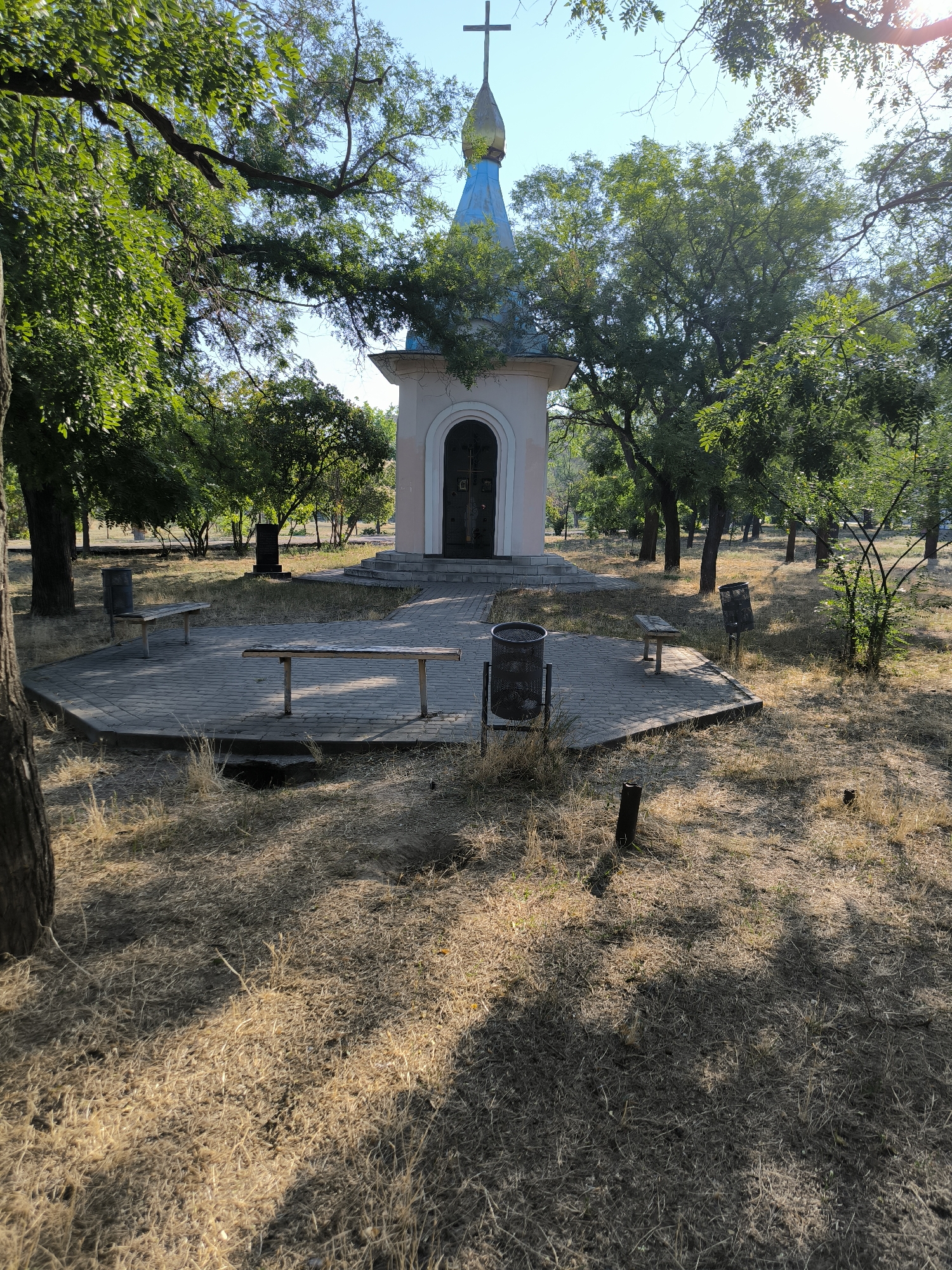 The image shows a small, light-pink chapel with a blue and gold domed roof and a cross on top, located in an outdoor, somewhat overgrown area. In front of the chapel, there is a circular paved area with several wooden benches and trash cans. The ground around the paved area is covered in dry grass and scattered leaves. Several trees with green foliage surround the chapel and the paved area. The sky is visible and appears clear. In the background, there are more trees and what appears to be a dark stone monument or grave marker. The scene suggests a place of quiet contemplation or memorial within a park-like setting. This location is the Chapel of the Holy Great Martyr Catherine located in the Victory Park of Mykolaiv, Ukraine.
