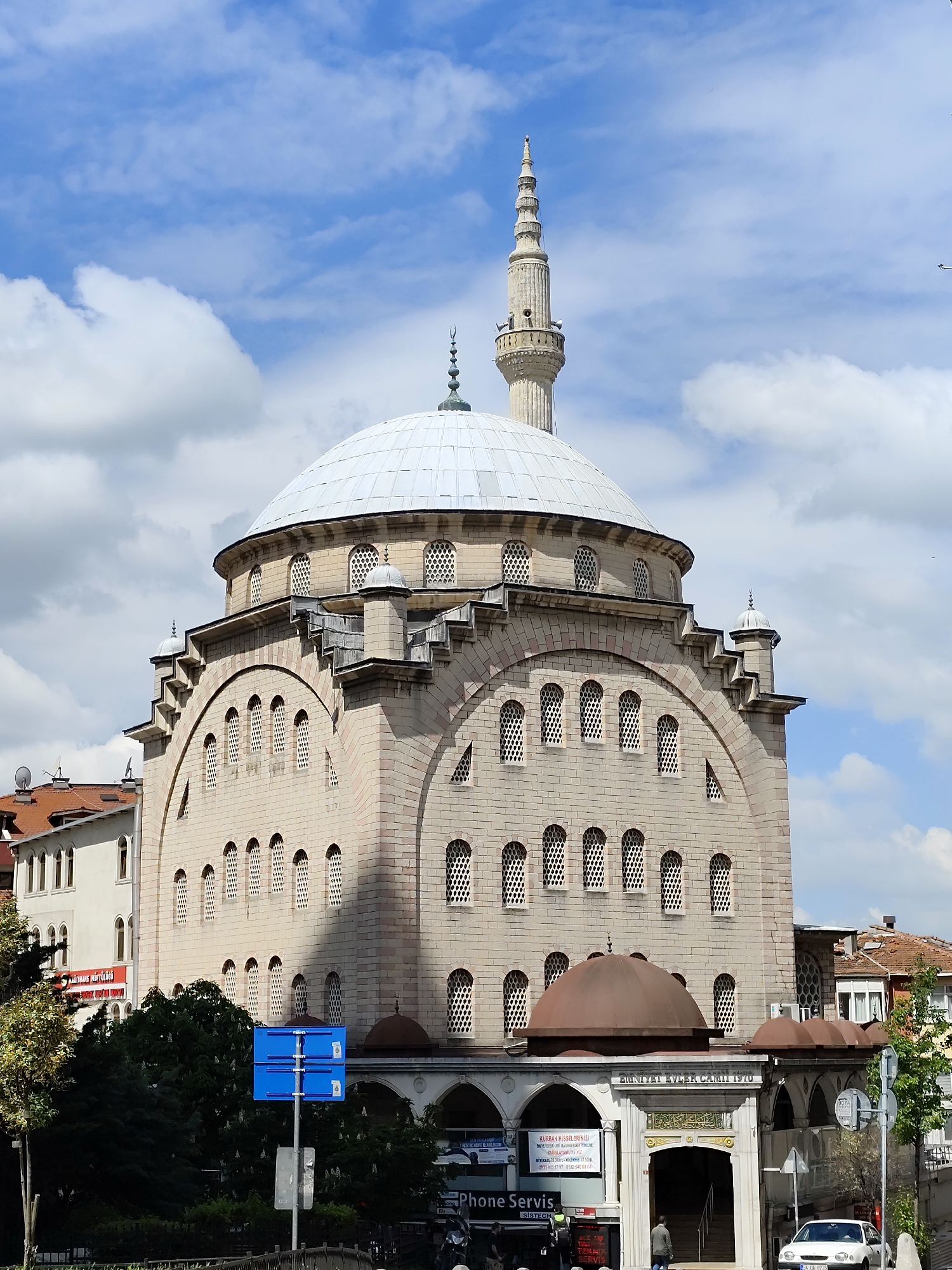 The image captures the exterior of the Zimniyat Evler Camii mosque, built in 1970, located in an urban setting, presumed to be in the city of Unknown. The mosque is characterized by its light-colored stone construction, large central dome with a white exterior, and a tall, slender minaret. Arched windows with lattice-like screens are visible on multiple levels of the building. A smaller, brown dome is situated near the base of the structure, over what appears to be an entrance area. Signage with Turkish script and Arabic calligraphy is visible over the entrance. A "Phone Servis" shop sign is placed on the entrance area. A pedestrian is visible walking toward the entrance of the mosque. In the foreground, there is a blue street sign and some green foliage. In the background, neighboring buildings with varying architectural styles are visible, along with a partly cloudy sky.