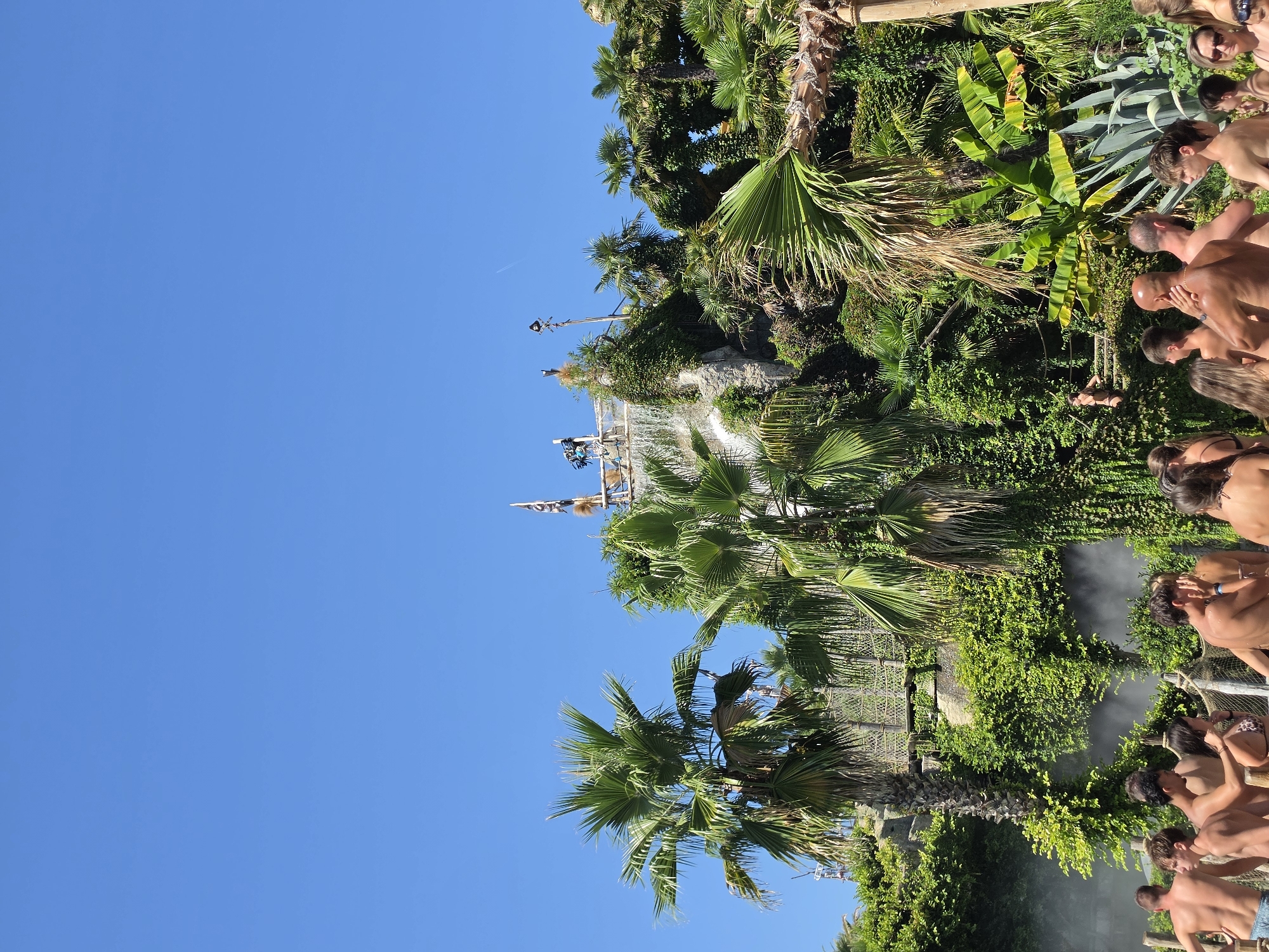 The image depicts a sunny day at a water park, likely Caribe Bay in Jesolo, Italy, with a clear blue sky dominating the upper portion. In the foreground, a group of people, mostly topless or wearing swimwear, are gathered, some appearing to be in a pool or waiting area, with mist rising around them. Palm trees and lush green vegetation are prominent, creating a tropical atmosphere. In the background, a man-made rock structure with a waterfall cascading down is visible, topped with wooden structures and flags, suggesting a themed area of the park. A rope bridge also spans across the rocks, further adding to the adventurous ambiance. The scene captures a moment of leisure and recreation within a vibrant, outdoor entertainment venue.