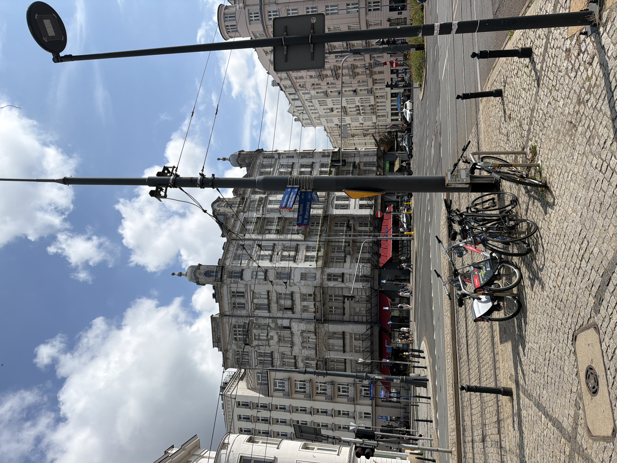 The image depicts a street intersection in Warsaw, Poland, likely near Plac Bankowy, characterized by historic, multi-story buildings with ornate facades and balconies. In the foreground, several bicycles are parked on a cobblestone pavement beside tram tracks. A prominent lamppost, a traffic signal, and street signs are visible, indicating a busy urban environment. People are seen walking on the sidewalks and crossing the street in the background, while cars navigate the intersection. The sky is partly cloudy, suggesting fair weather.