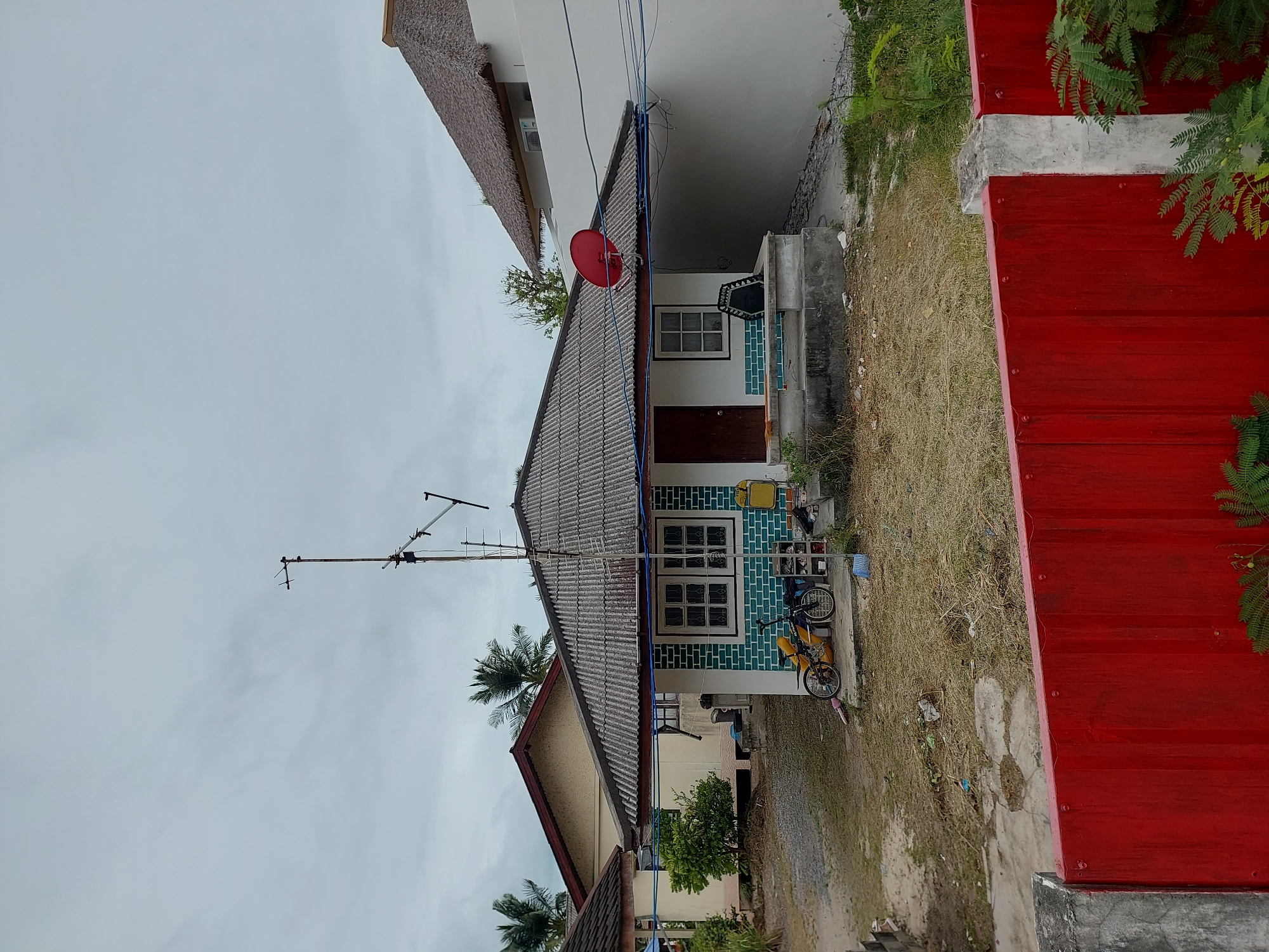 A single-story house with a grey tiled roof is situated in a residential area on Ko Samui District, Thailand. The house has white walls with a section of teal brickwork around the windows. A satellite dish is mounted on the roof, and an antenna is attached to the side of the house. In front of the house, there is a small yard with dry grass and some debris. A yellow bicycle is leaning against the teal brickwork. To the right of the house, a white wall with a thatched-roof structure is visible. A bright red metal fence runs across the foreground of the image. The sky is overcast.
