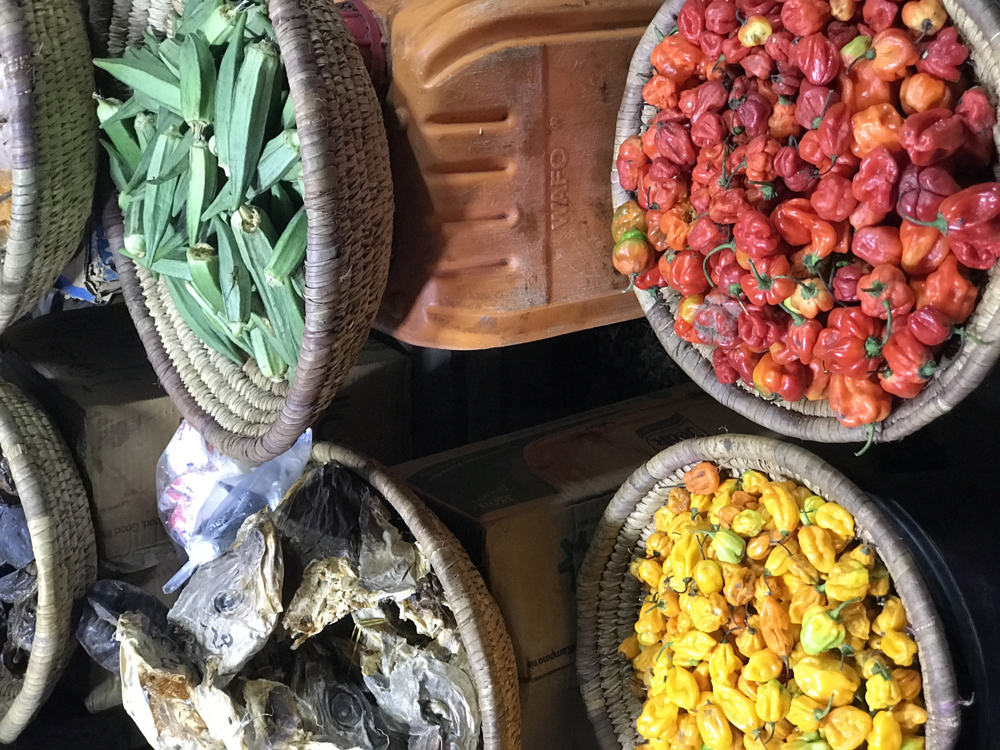 An overhead view captures a vibrant display of fresh produce and dried goods arranged in traditional woven baskets, likely within a market setting in Abuja, Nigeria. The scene is focused solely on the items, with no people visible.
In the top left, a light brown woven basket is filled to the brim with numerous fresh, green okra pods. Adjacent to it, centrally positioned, is a sturdy, orange-brown plastic container, possibly a bucket or crate, with the word "WAFO" visibly embossed on its surface.
To the top right, another woven basket holds a generous quantity of small, intensely colored chili peppers, primarily a mix of bright red and orange hues, characteristic of varieties like Scotch bonnets or Habaneros.
At the bottom left, a third basket contains pieces of dried fish, including several distinct fish heads, suggesting a common method of food preservation. Surrounding this basket are glimpses of plastic bags and cardboard boxes, one of which shows partially visible text, including "Good li" (likely "Good life").
Completing the arrangement at the bottom right is a fourth woven basket, overflowing with an array of vibrant yellow chili peppers, similar in shape and size to the red ones. Nearby, another cardboard box reveals text fragments such as "POSE" and "campigna le." The overall impression is one of abundant provisions and local commerce.