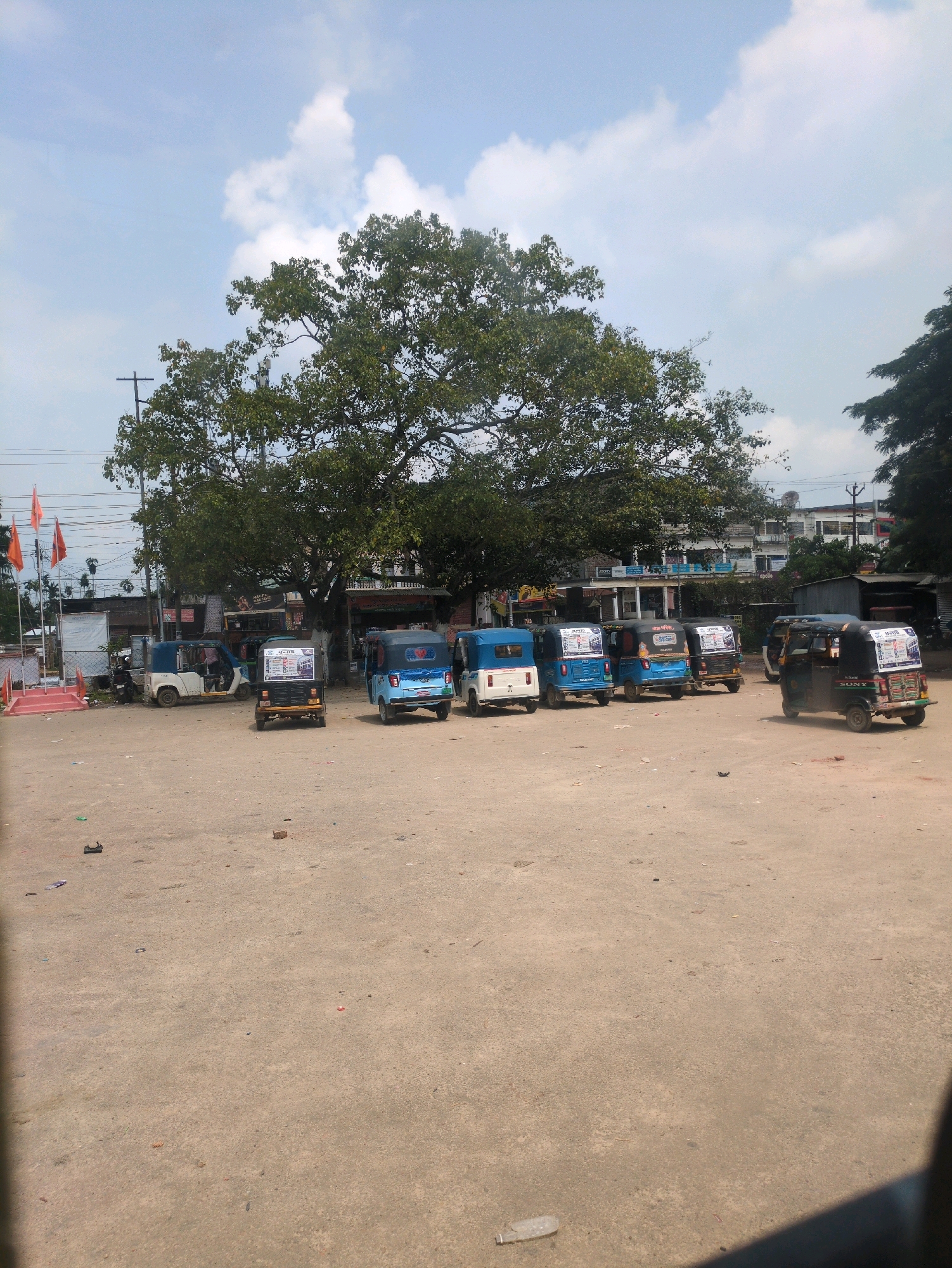 The image captures a scene in the city of Ambassa, India, featuring a parked collection of auto-rickshaws on a dusty, open lot. The sky above is partly cloudy with a bright, clear blue expanse. Dominating the background is a large, mature tree with dense green foliage, providing shade over the area. Behind the tree, several low-rise buildings are visible, some appearing to be shops or residences, with a satellite dish perched atop one.
In the foreground and mid-ground, an array of auto-rickshaws are parked in rows. Several are blue, while others are white or yellow, and one prominently displays advertising. The ground is a rough, dirt surface with scattered debris, including small stones, a plastic bottle, and cigarette butts.
On the far left of the image, a small, elevated platform with orange flags on poles is present, suggesting a public space or an area of significance. Other parked vehicles, including a motorcycle, are also visible. The overall impression is that of a transportation hub or a busy roadside area in a small Indian city.