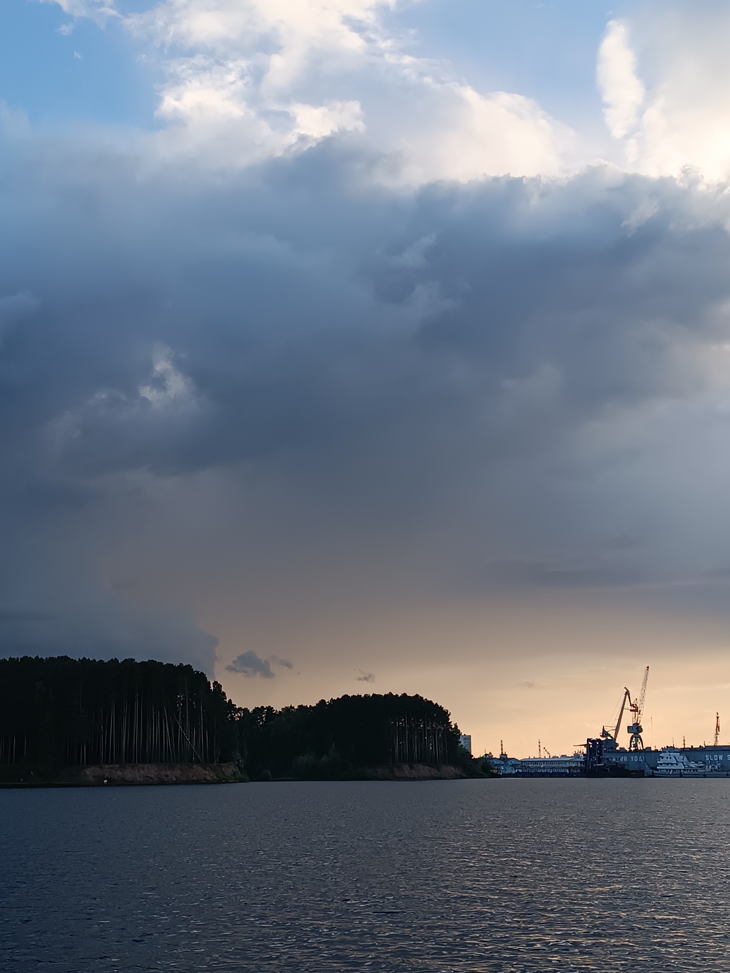 The image depicts a body of water in the city of Chaikovsky, Russia, under a dramatic sky. On the left side, a dense forest lines the shore, with tall, thin trees. A sandy embankment is visible beneath the trees. The water itself is dark and slightly rippled.
On the right side of the image, a port area is visible. Several ships are docked, and large cranes, characteristic of a port or shipyard, dominate the skyline. The sky above is a mix of dark, heavy clouds and lighter, brighter areas, suggesting either the aftermath of a storm or the approach of sunset. The overall mood is atmospheric and somewhat dramatic due to the lighting and cloud formations. No people are clearly visible in the scene.