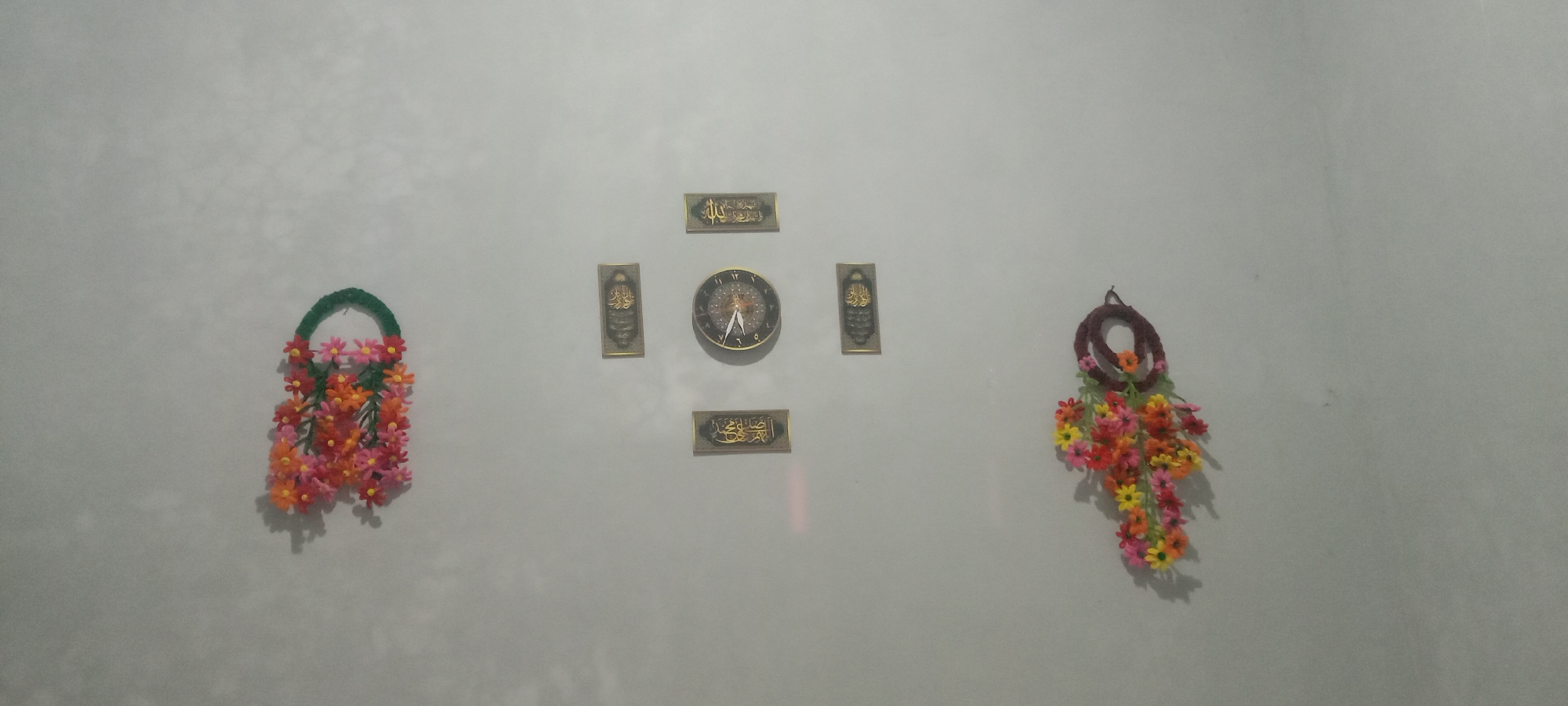 The image shows a wall with several decorative items. On the left, there is a green basket-shaped decoration with an arrangement of colorful flowers, predominantly pink, orange, and yellow. In the center of the wall, there is a clock with a dark brown frame and a clock face displaying Roman numerals. Above and below the clock are two rectangular plaques with Arabic calligraphy. To the right of the clock, there is another vertical rectangular plaque with Arabic calligraphy. On the far right, there is a similar decorative item to the one on the left, consisting of a circular braided frame with an arrangement of colorful flowers hanging from it, featuring pink, orange, and yellow hues. The wall itself appears to be a light, textured gray. The scene is indoors, likely a home or office, given the decorative nature of the objects. There are no people visible in the image. The lighting suggests it might be evening or a dimly lit room, with shadows cast by the objects onto the wall. The image does not provide any specific information about the location within Utama Jaya Mataram, Indonesia, beyond what can be inferred from the style of the decorations, which are common in many parts of Southeast Asia.