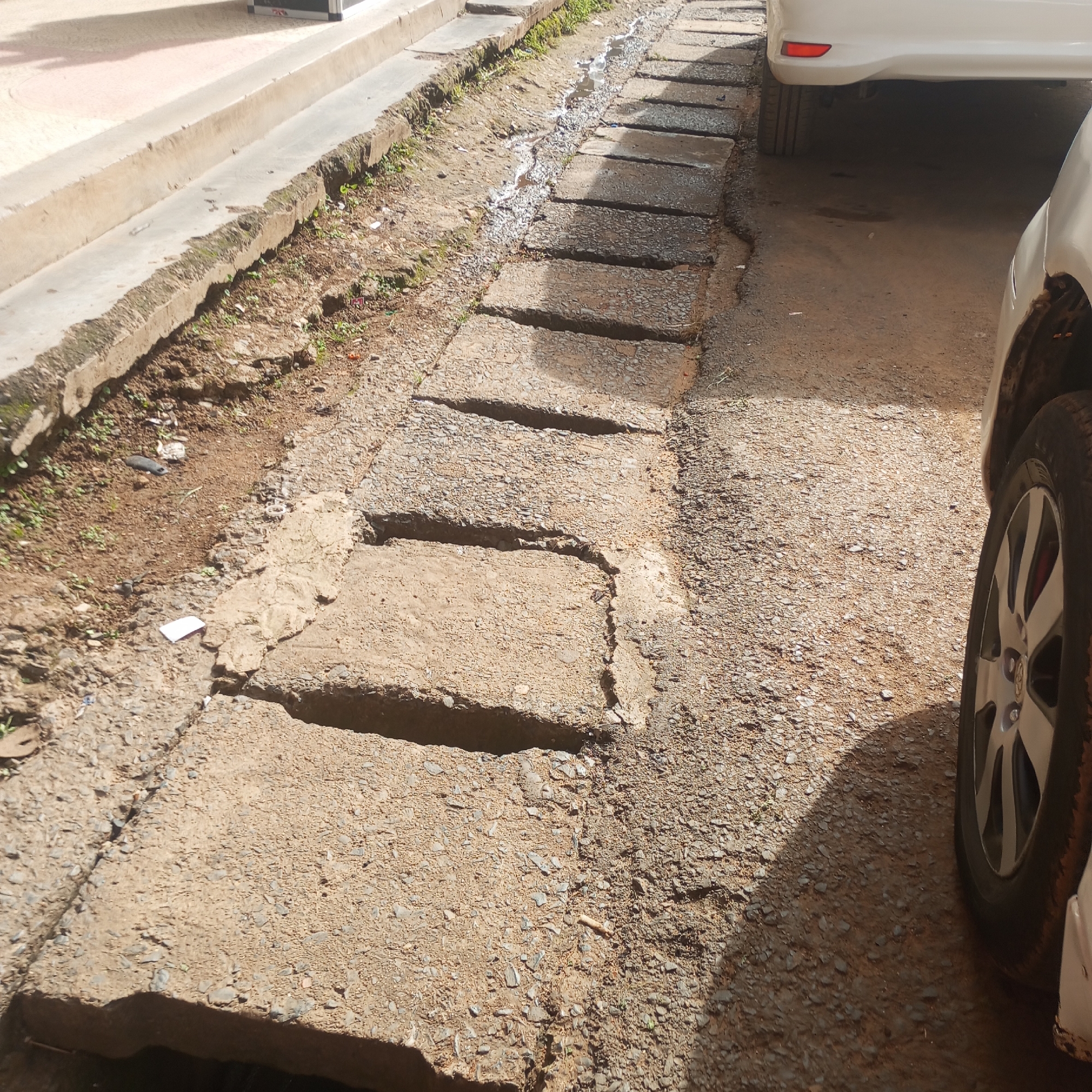 The image depicts a deteriorated drainage system in Sheywe ward, Kenya. The system consists of concrete slabs covering a drainage channel. The slabs are cracked, broken, and misaligned. There is stagnant water and debris visible within the channel. Adjacent to the drainage system is a dirt road surface comprised of loose gravel. Two white cars are partially visible, one with a tire directly beside the drain. Debris such as pieces of paper are visible on the roadside. A concrete curb for a building is visible next to the dirt verge beside the drainage system. The overall impression is one of neglect and disrepair.