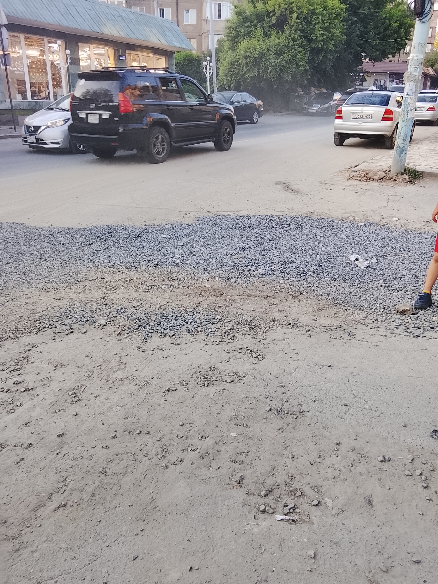 The image captures an outdoor scene on a street that appears to be under construction or in disrepair. In the foreground, a patch of gravel lies on the dirt ground, suggesting roadwork. Further into the image, a dark-colored SUV is parked on the asphalt road, and a silver sedan is driving away. Behind the vehicles, a line of trees creates a backdrop, and buildings are visible in the distance. The overall impression is one of an ordinary day in a somewhat unfinished urban environment. The lighting suggests it is daytime, possibly late afternoon. There is no significant weather activity. The only visible text is on the license plate of the silver sedan, which reads "36 CM 411". The scene is devoid of any strong emotional cues, presenting a straightforward depiction of a street with passing vehicles and ongoing urban development.