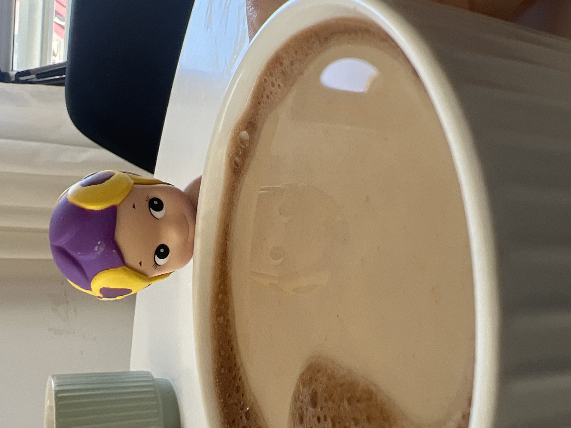 A close-up, vertically oriented image captures a whimsical indoor scene featuring a white ceramic mug filled with a light brown, frothy beverage, and a small toy figure. The mug, occupying the right and upper portions of the frame, contains a drink resembling coffee or hot chocolate, topped with a thick layer of light foam. Darker, bubbling froth adheres to the inner rim of the mug.
To the left of the mug, a toy figurine, appearing to be a girl, peeks playfully into the drink. The toy has a light-colored face with large, expressive black eyes and long eyelashes, looking upward. Her head is adorned with molded purple hair or a cap, detailed with yellow accents. A small, light green cylindrical object with a ribbed texture is partially visible in the bottom-left corner.
The background includes a soft, bright light source, likely a window, visible in the upper-left, and a dark, indistinct object (possibly furniture or a curtain) extending across the upper part of the frame. The overall setting is bright and clean, with white surfaces dominating. The image conveys a sense of playful curiosity. No text is visible in the frame. The location context for this indoor scene is Budva, Montenegro.