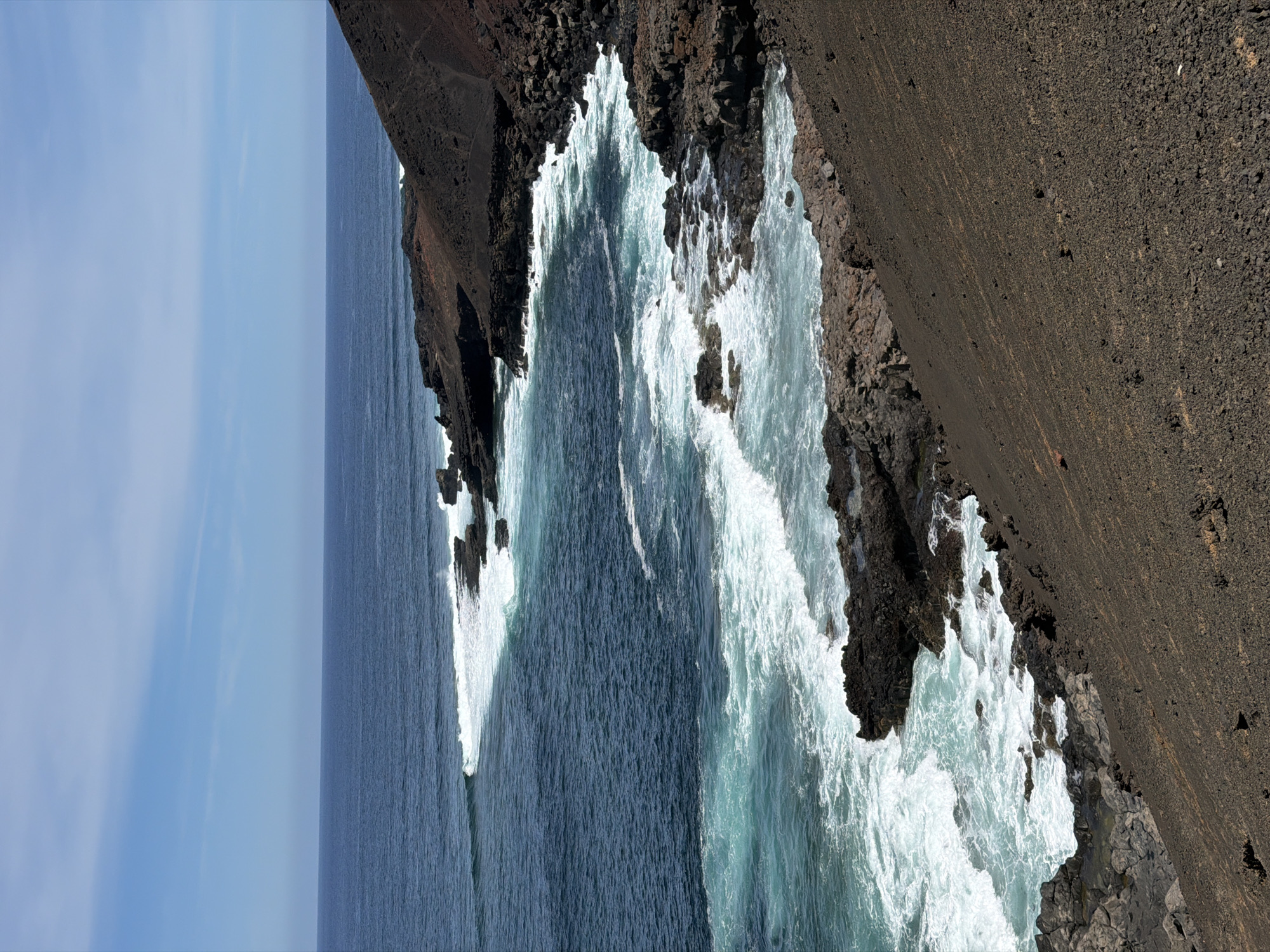 The image captures a coastal scene in Tinajo, Spain. The composition showcases a rugged, dark volcanic coastline being battered by ocean waves. Whitewater foam contrasts sharply with the deep blue of the sea and the black rocks. The sky is visible as a strip of blue at the edge. No people or other subjects are visible. The primary action is the waves crashing against the rocky shore.