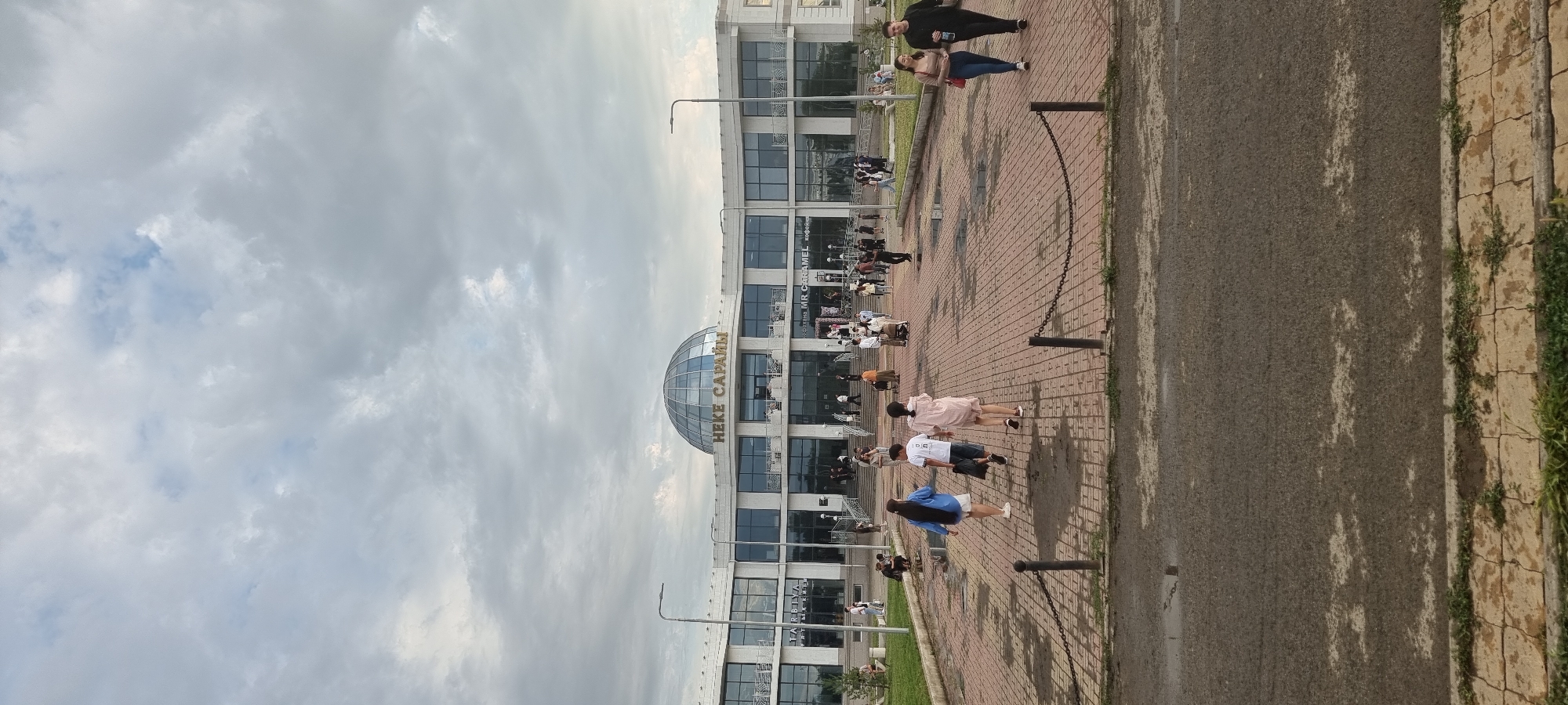 The image depicts the exterior of the "NEKE SARAI" building, a prominent landmark in Aktobe, Kazakhstan. The building features a modern architectural design with large glass windows and a distinctive dome. In front of the building, a paved plaza area is visible, with several people walking and gathered. The plaza is bordered by planters and a walkway. The sky is overcast with clouds, suggesting a temperate weather condition. The overall scene conveys a public space with moderate foot traffic.